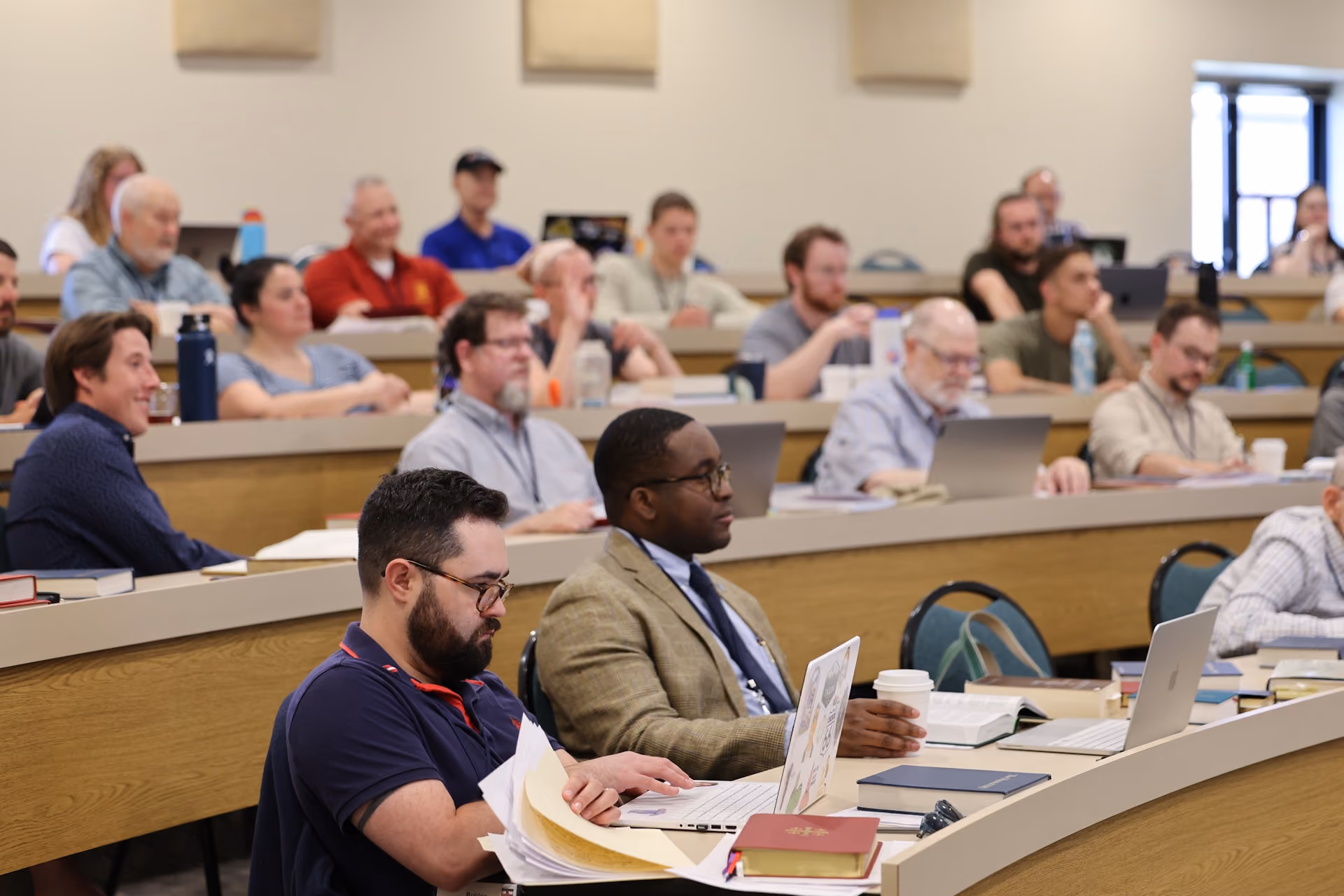 Adults seated in a lecture hall attending a class or seminar with laptops, books, and notes.