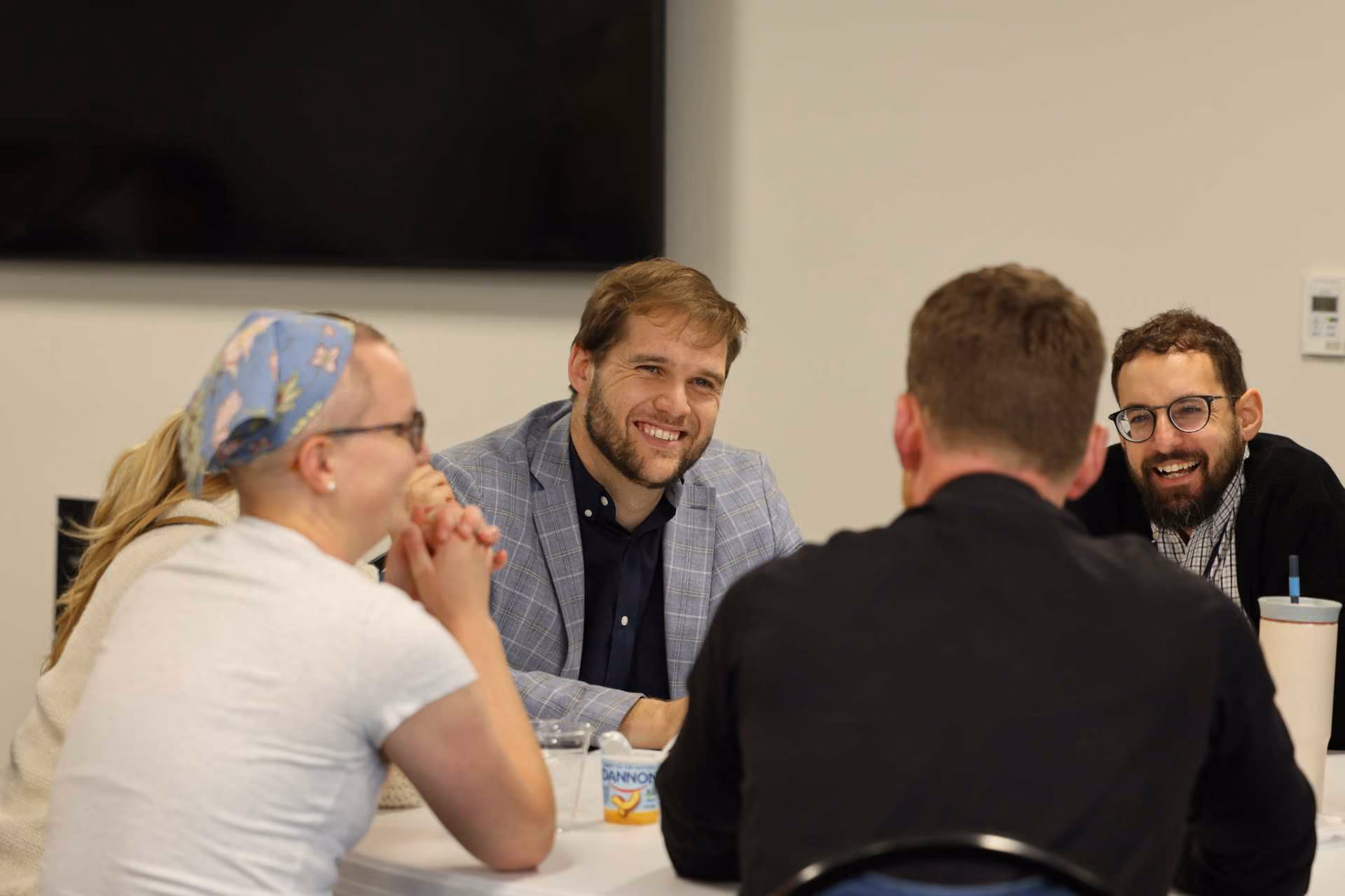 Four people are seated around a table in a bright classroom setting, smiling and engaged in conversation. A wall-mounted screen is visible in the background.