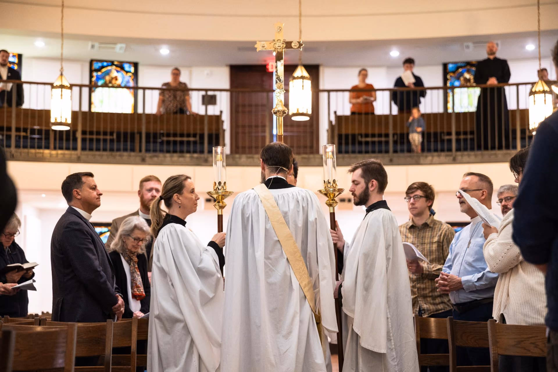 Two acolytes carrying tall candlesticks flank a robed minister leading a processional cross down the center aisle, while worshippers stand in pews on the main floor and a balcony above.