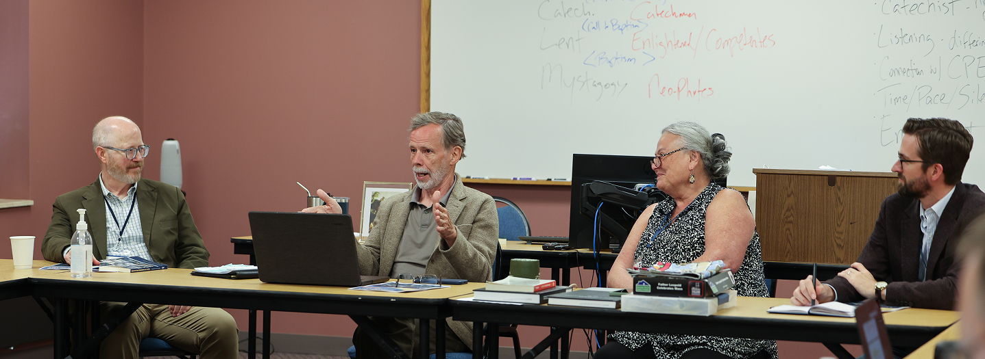 Four people sit at tables arranged in a panel format in a seminary classroom. One man gestures as he speaks while the others listen; a whiteboard behind them shows notes on catechesis stages including pre-catechumenate, catechesis, and mystagogy.