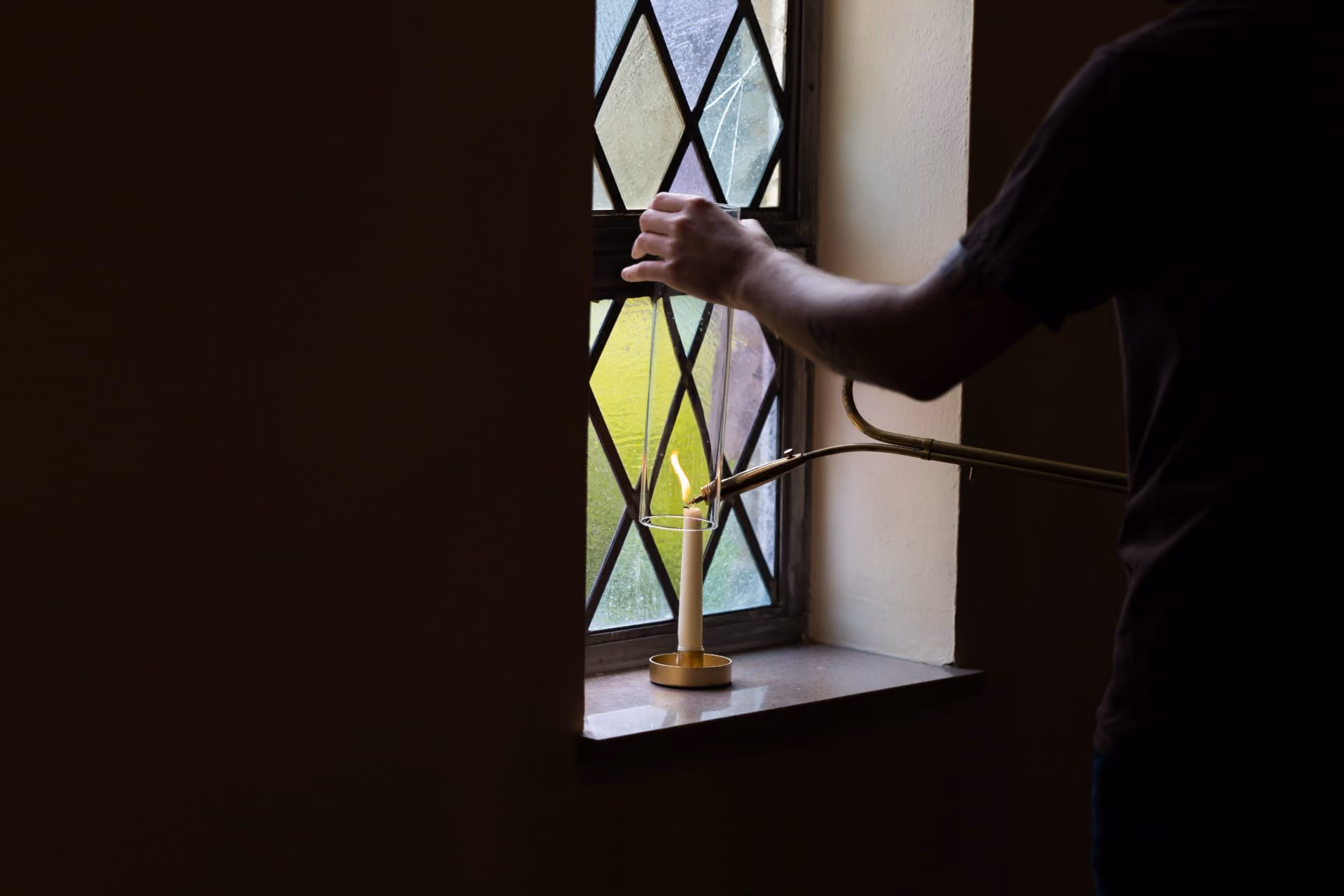 Person lighting a candle on a window ledge with an oil lamp through a stained glass window.