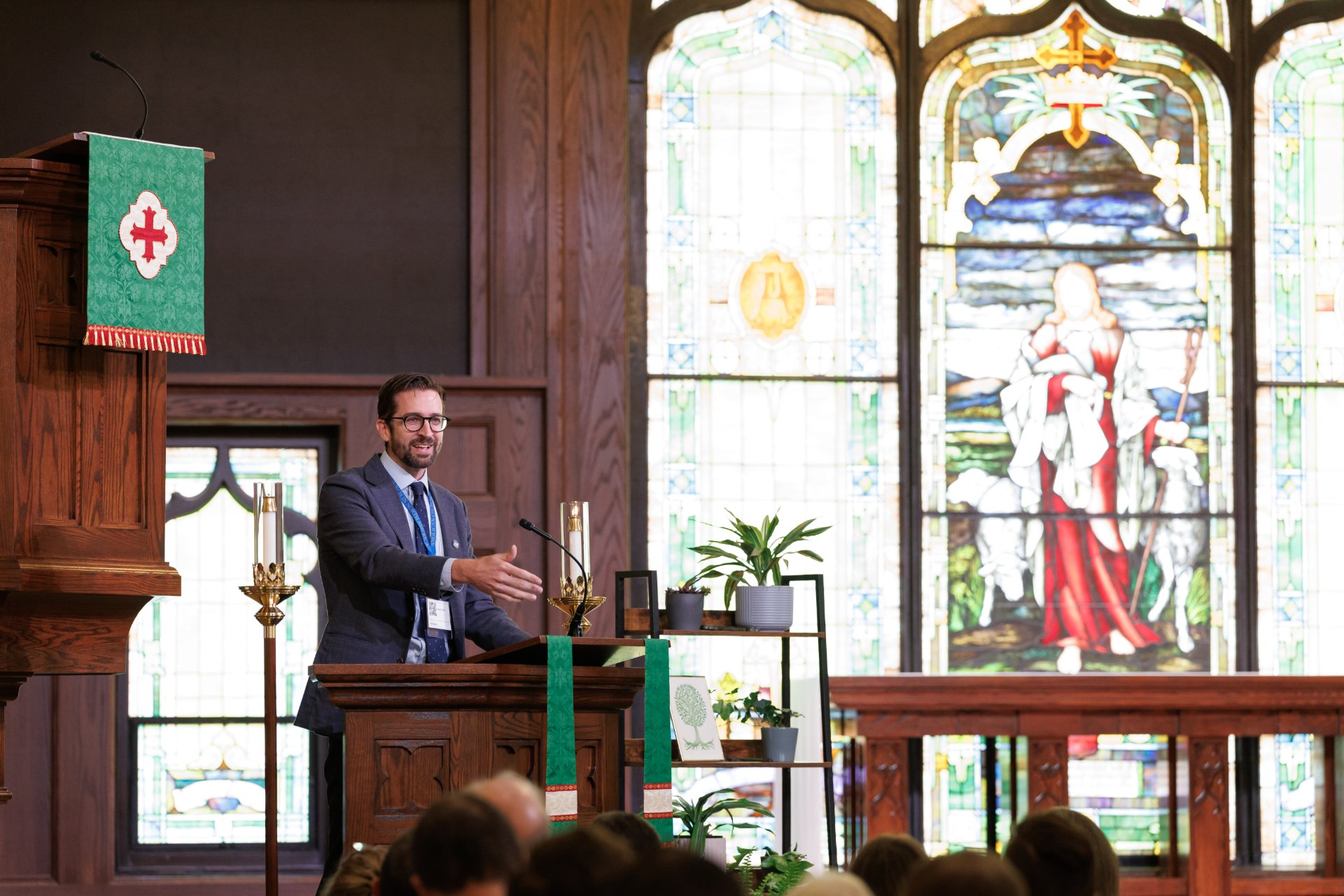 A man in a suit and lanyard gestures while speaking at a wooden pulpit in an Anglican church. A green parament with a cross hangs nearby, and a large stained glass window depicting the Good Shepherd fills the background.