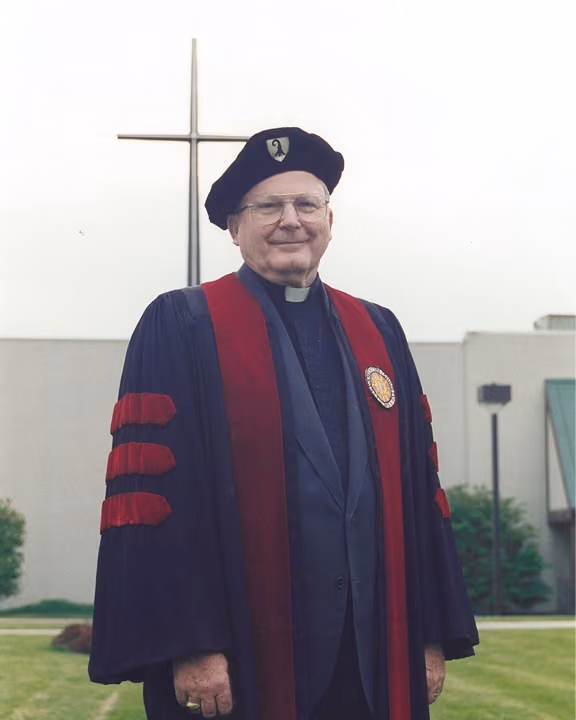 An elderly man wearing academic regalia and a clerical collar stands outdoors with a cross in the background.