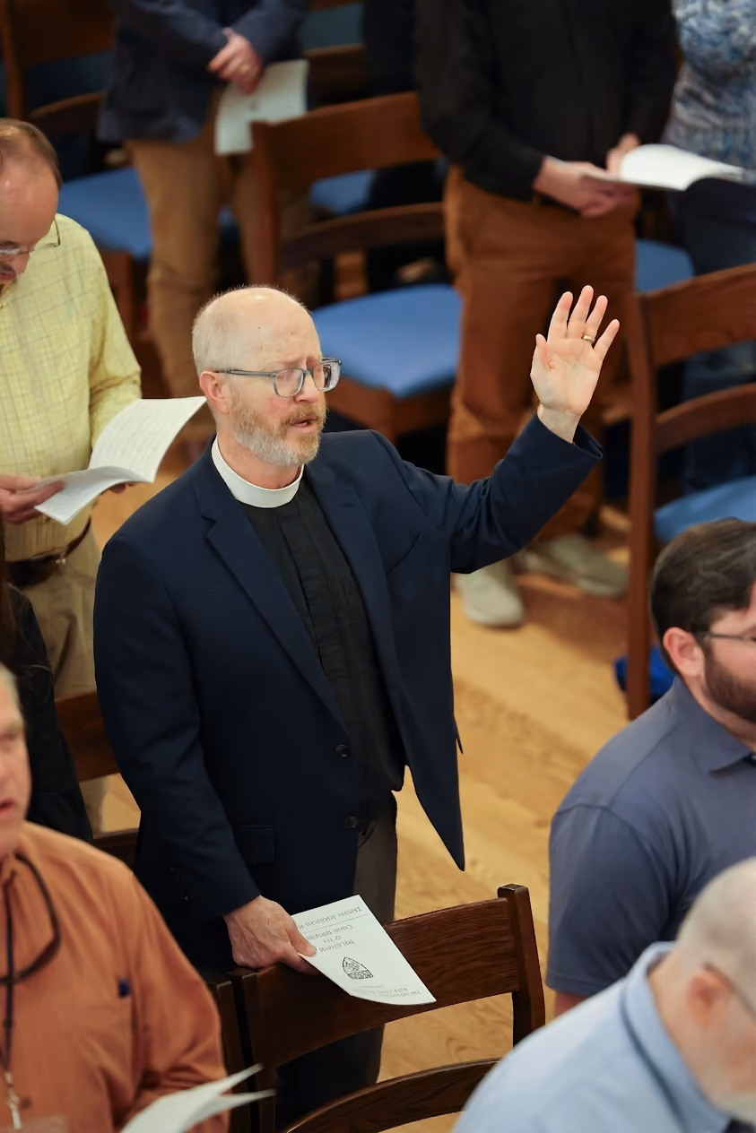 Clergyman with glasses raising hand holding a paper in a seated congregation during a service.
