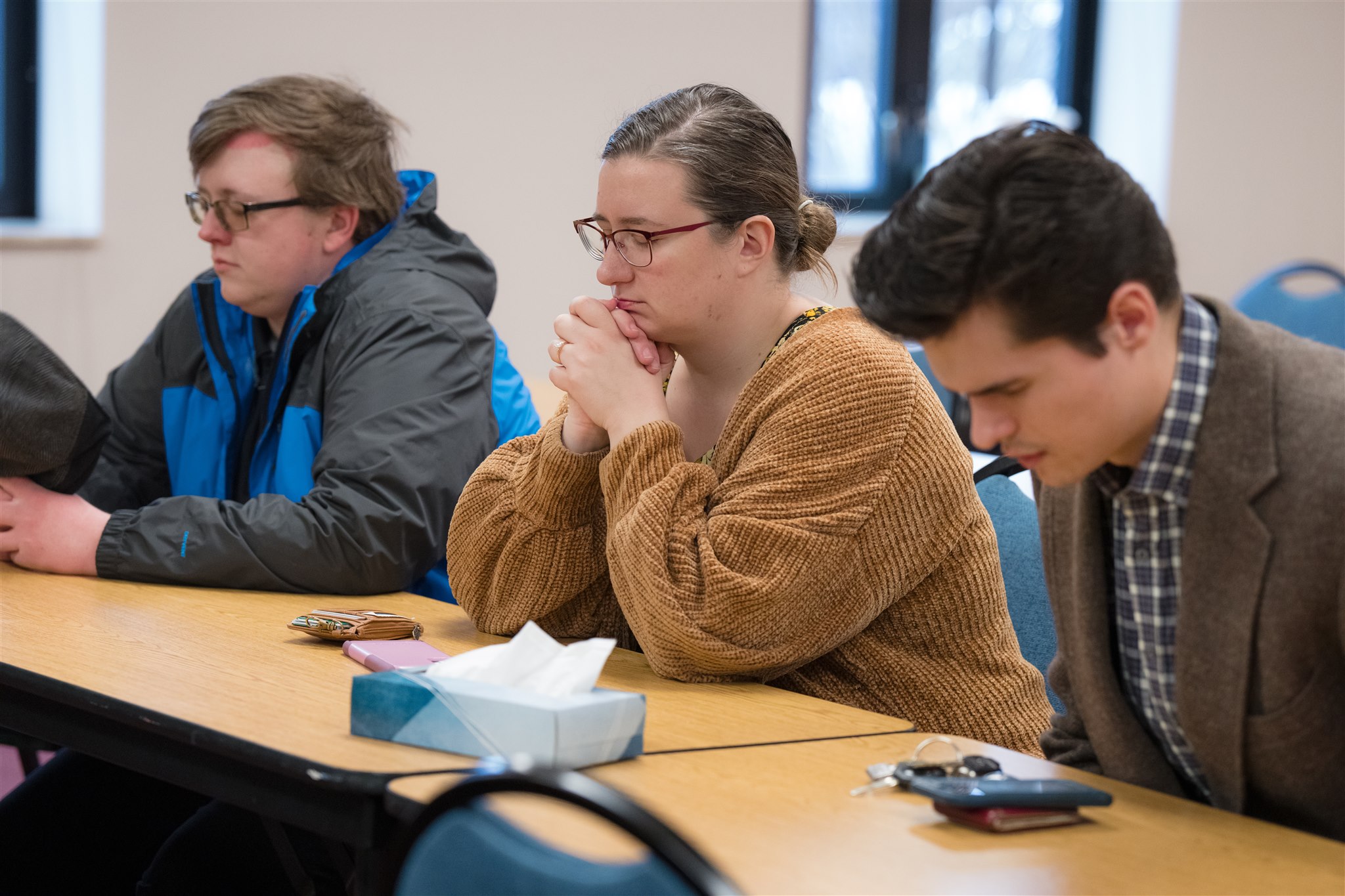 Three people sit solemnly at a table in a room, hands clasped, appearing deep in thought. A tissue box and personal items lie on the table.