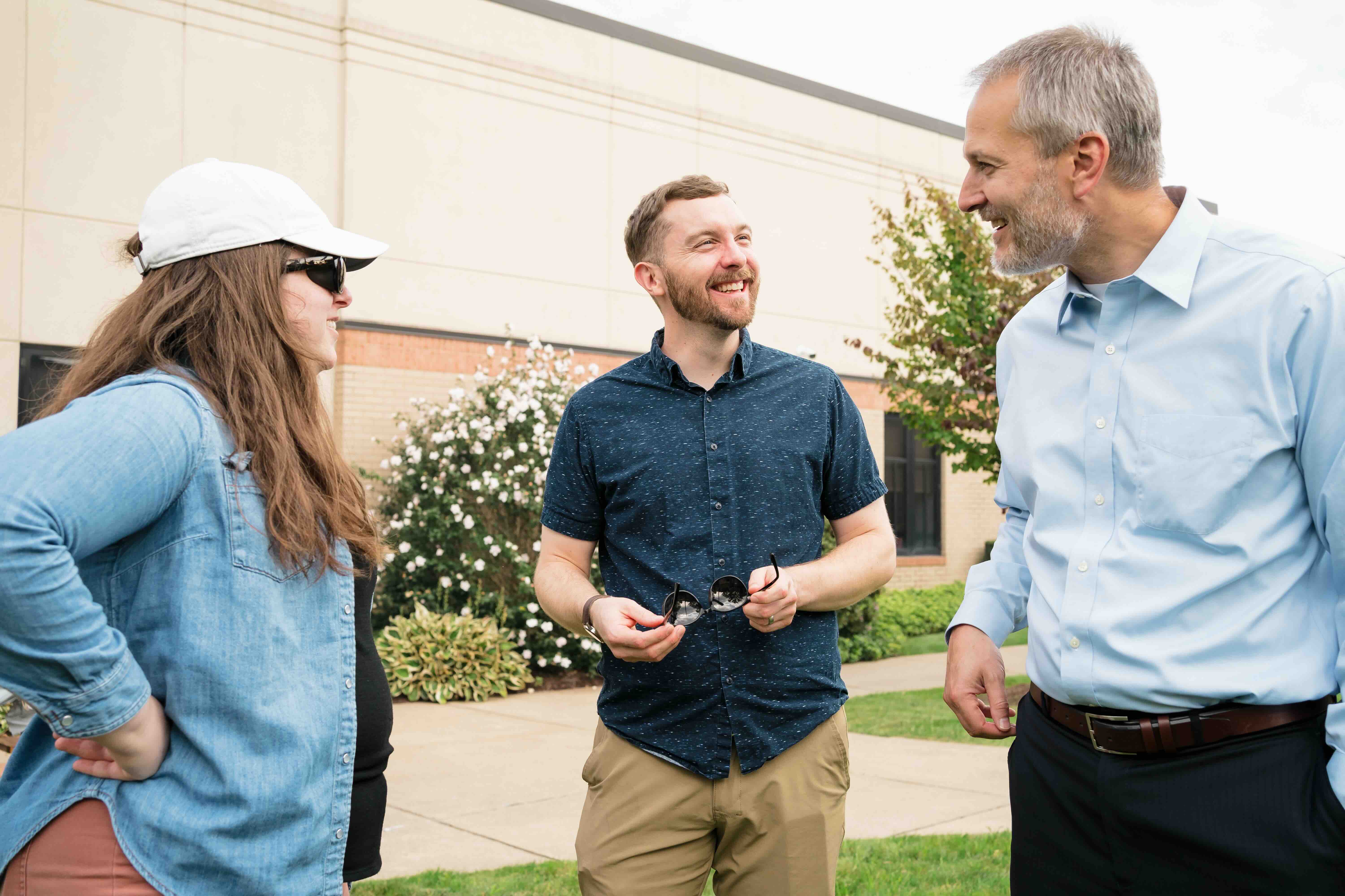 Three people stand outside, engaged in a lively conversation. The atmosphere is friendly and relaxed. A woman in a cap and two men smile and chat.