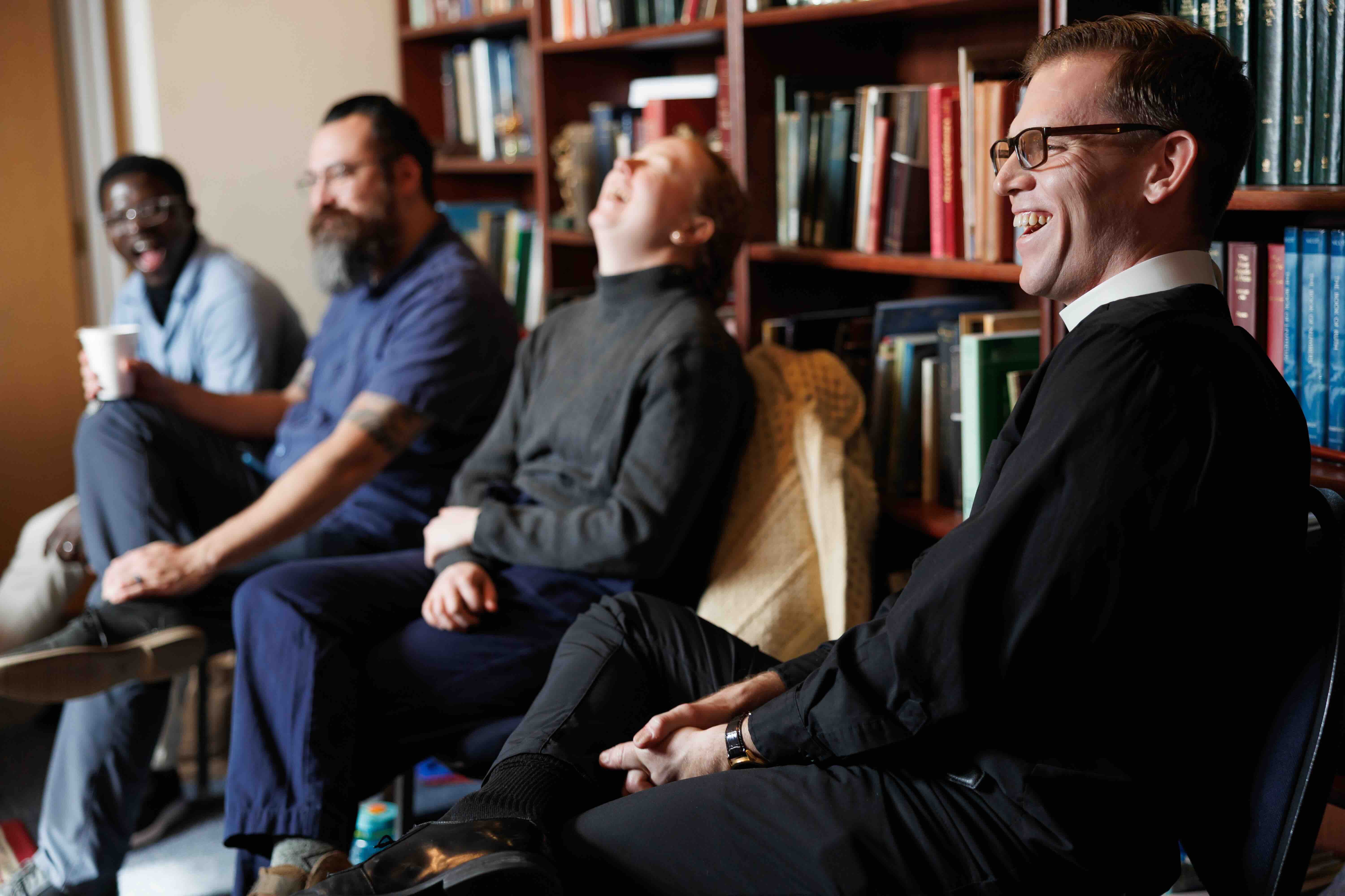 A group of four people sits in a library, laughing and smiling. Books fill the shelves behind them, creating a warm, relaxed atmosphere.