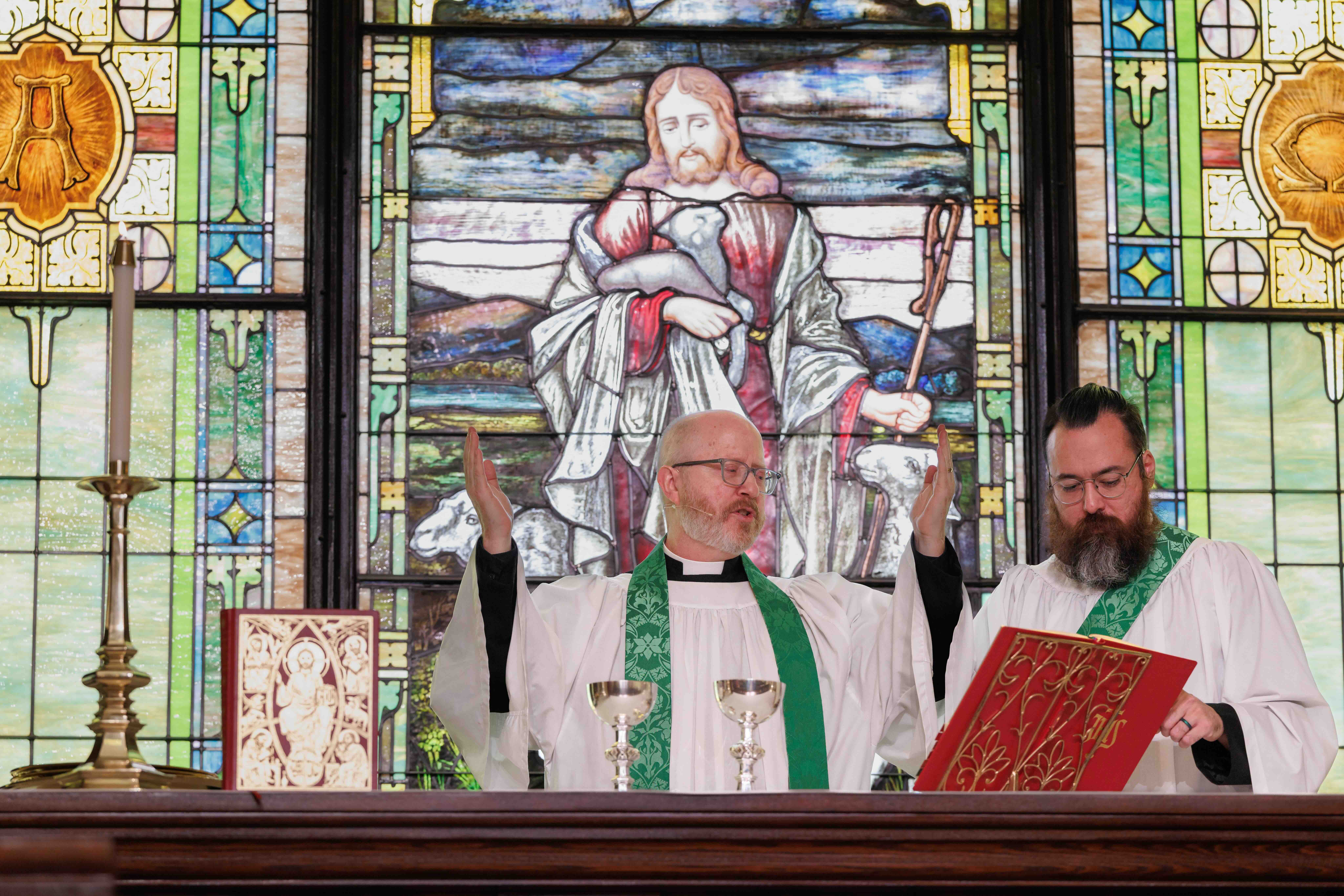 Two clergy members in green stoles lead a church service at the altar, with a stained glass window of Jesus holding a lamb in the background. The atmosphere is solemn and reverent.