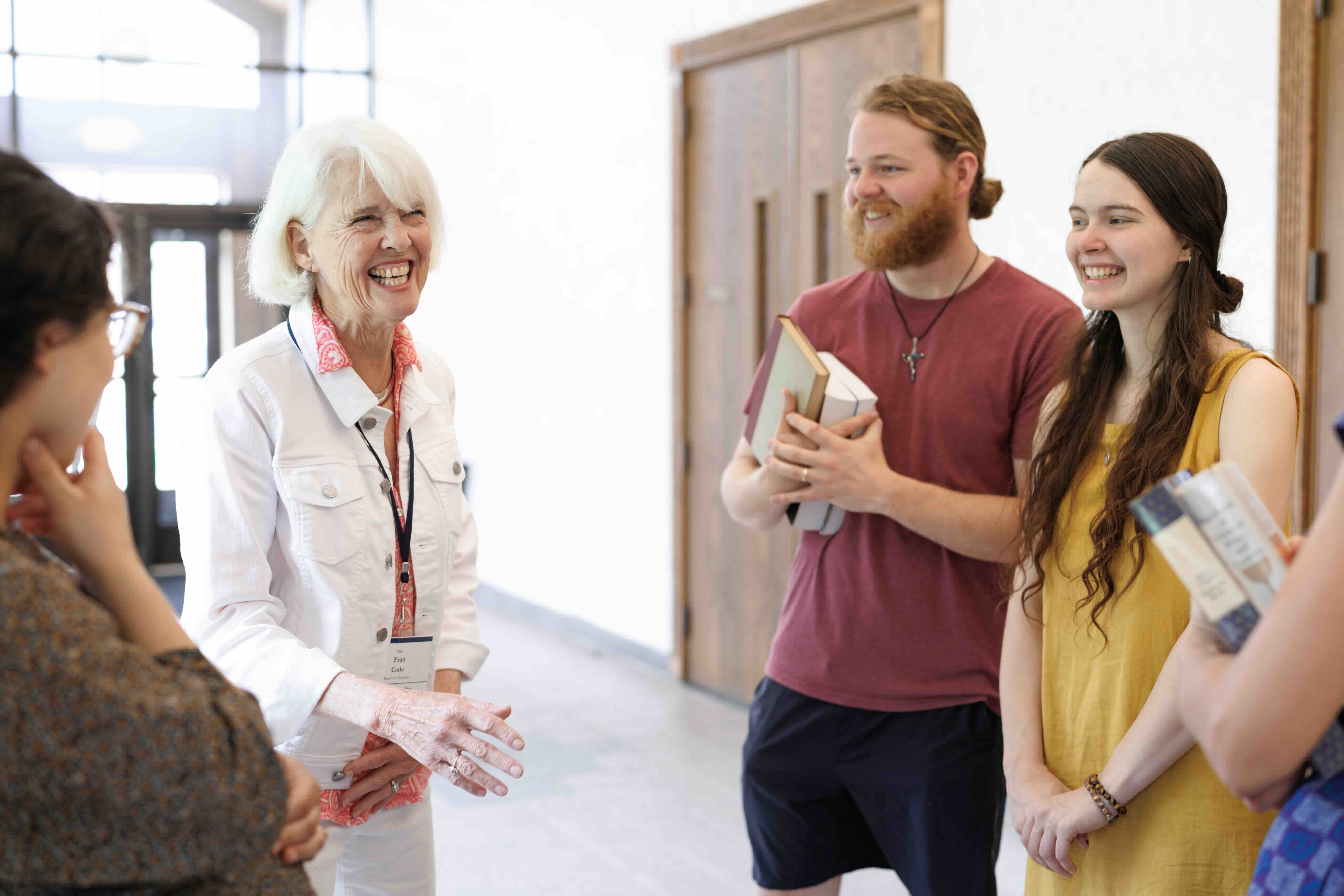 A diverse group of people is joyfully conversing in a bright hallway. An older woman in a white jacket smiles warmly, as others hold books, conveying a sense of camaraderie and learning.