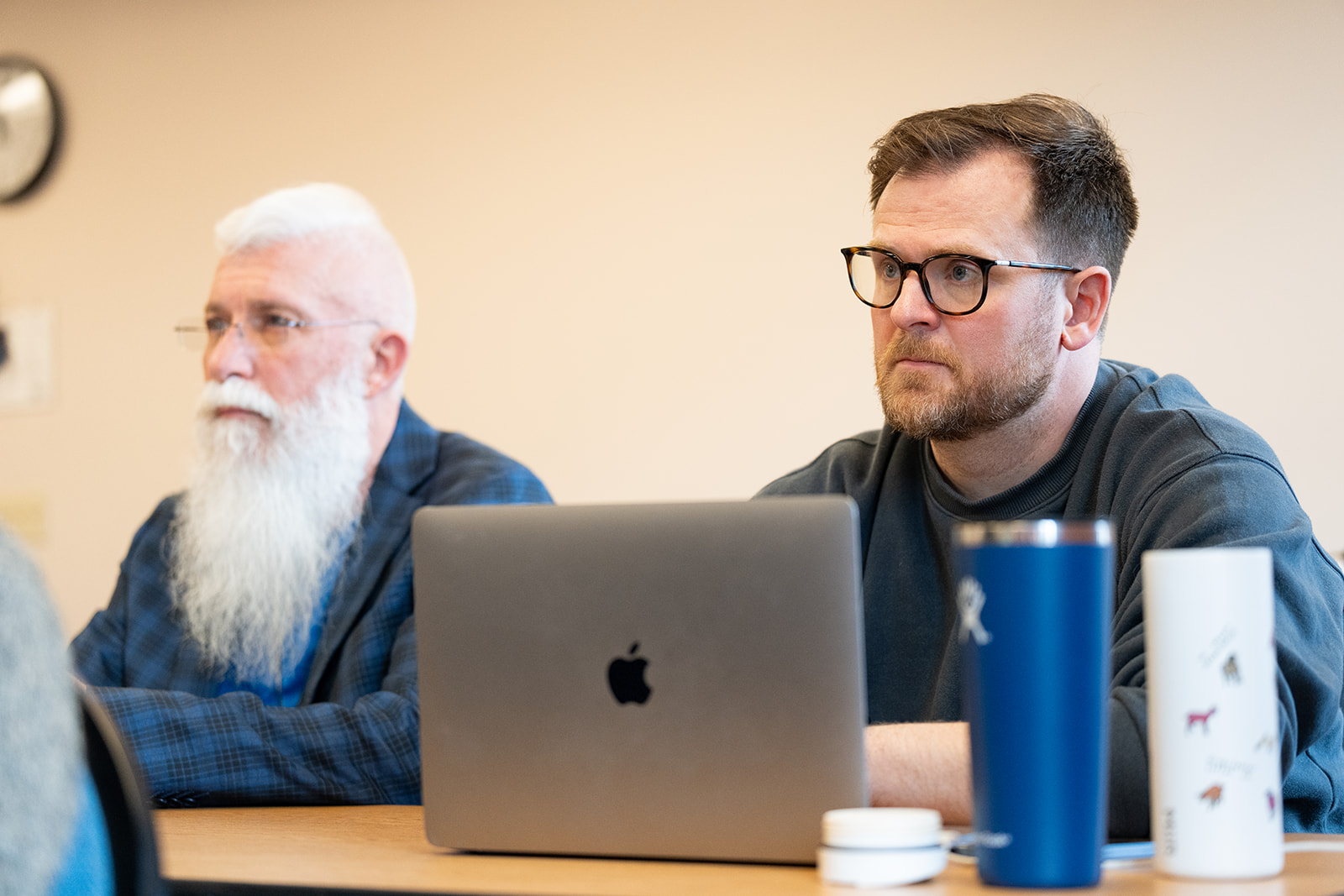 Two men sitting at a table, focused on a meeting. One has a white beard and glasses; the other wears glasses and sits behind a laptop.
