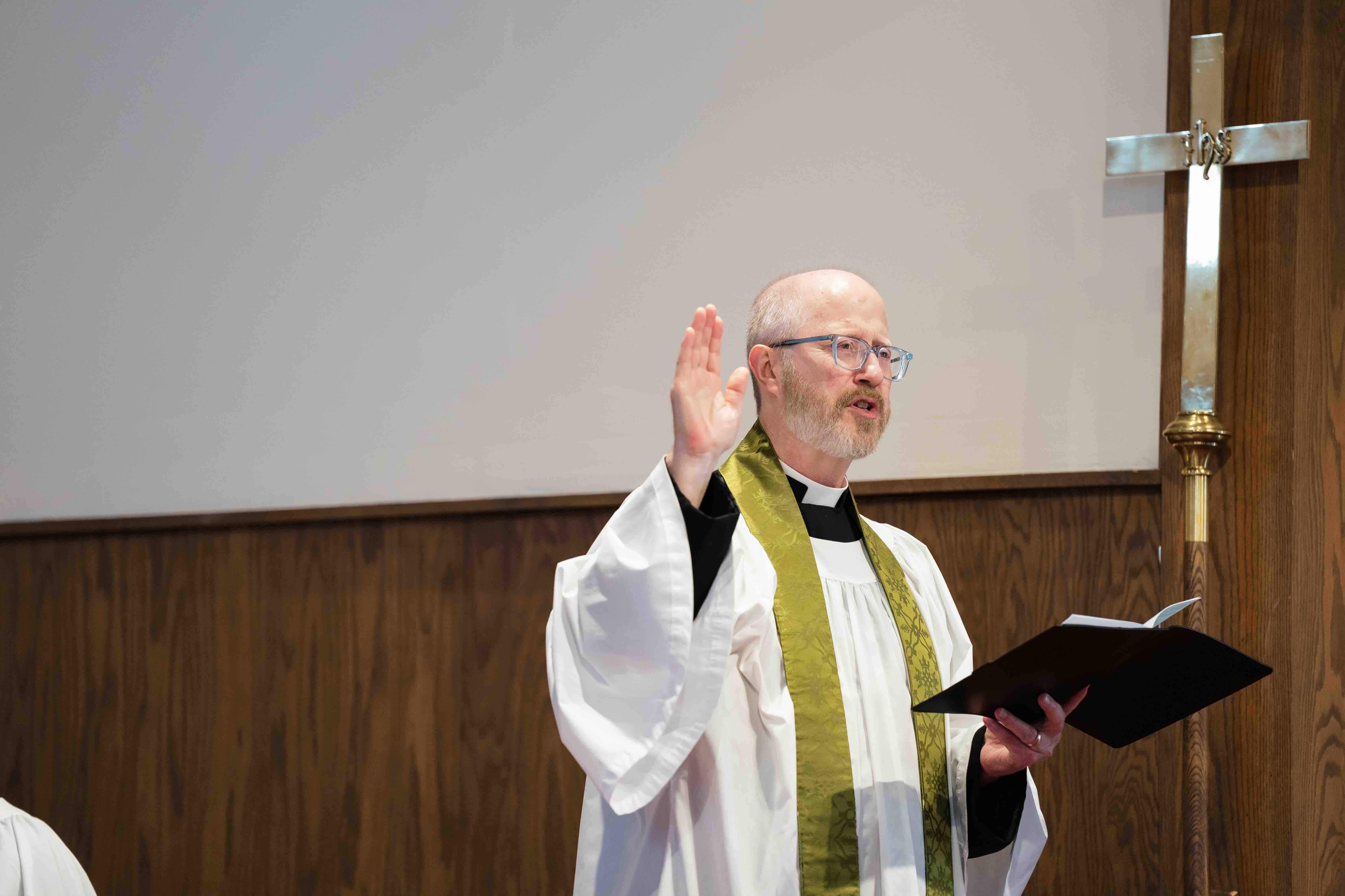 A priest in white robes with a green stole gestures while holding an open book, standing by a wooden cross in a church setting, conveying a solemn tone.