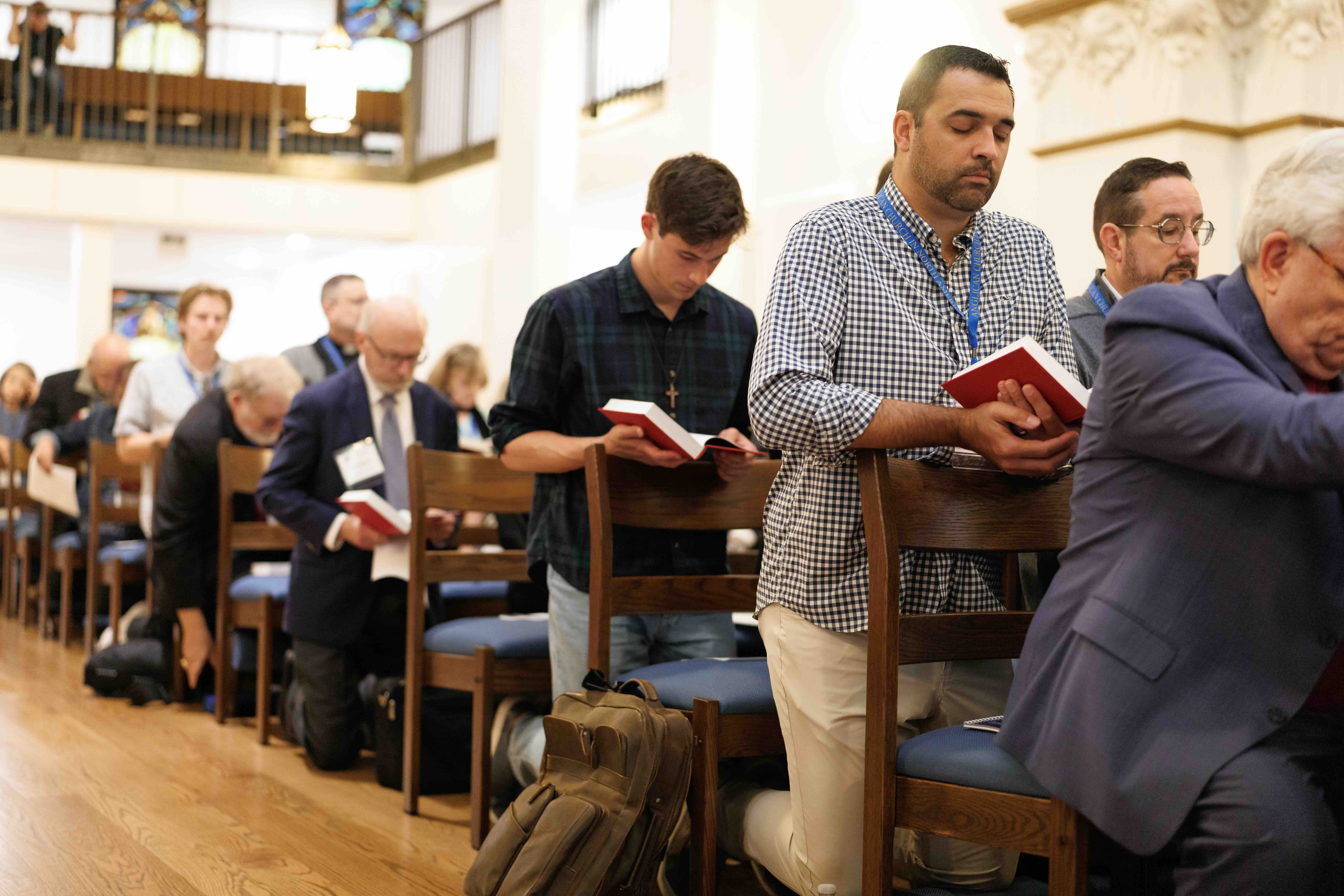 A group of people kneeling in prayer inside a church, each holding a red book. The scene conveys solemnity and devotion, with wooden pews and soft lighting.