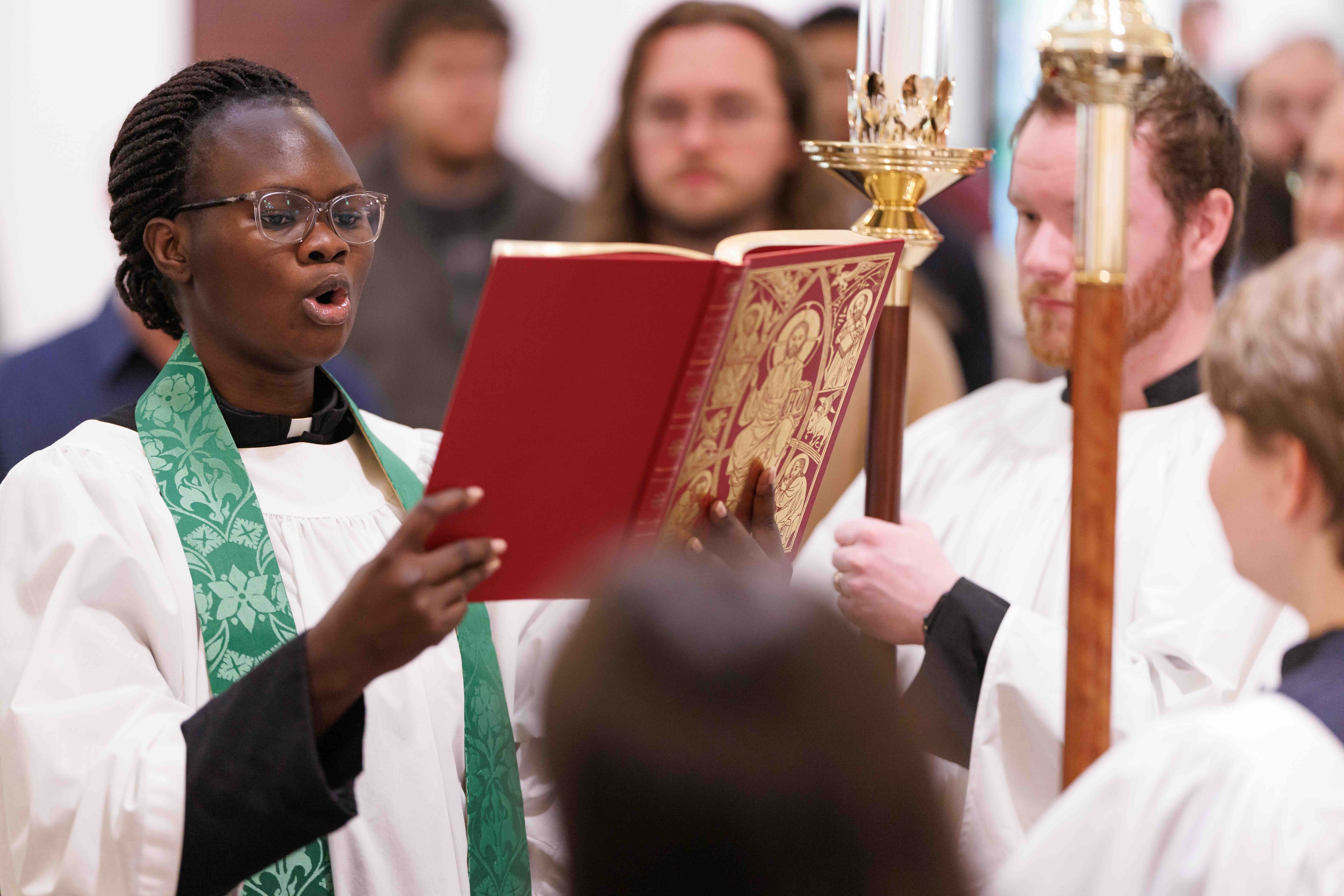 A person in religious robes sings from an ornate red book, surrounded by others in white vestments, conveying a solemn and reverent atmosphere.
