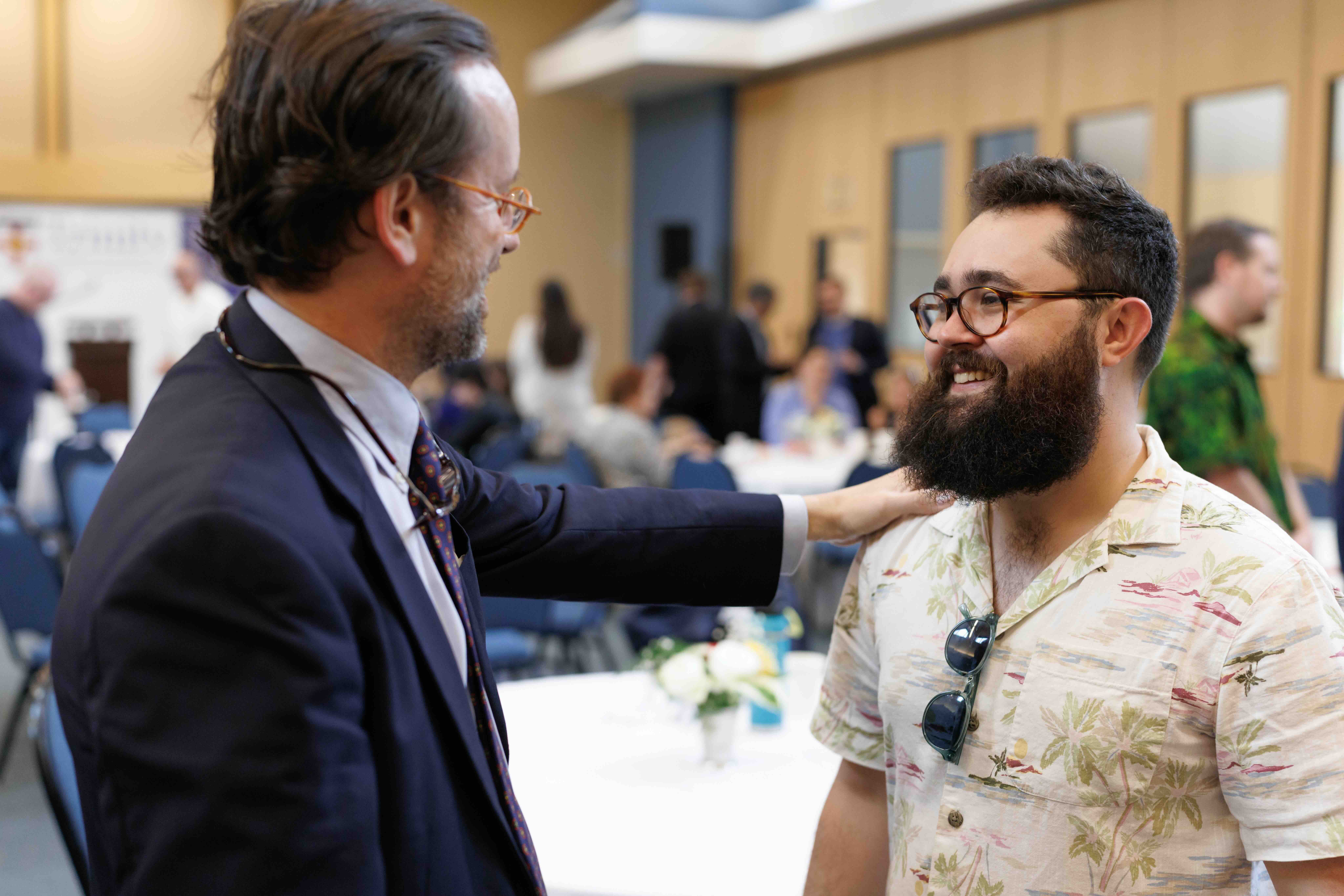 A man in a suit and glasses warmly greets another smiling man in a floral shirt at a social event, conveying a friendly and welcoming atmosphere.