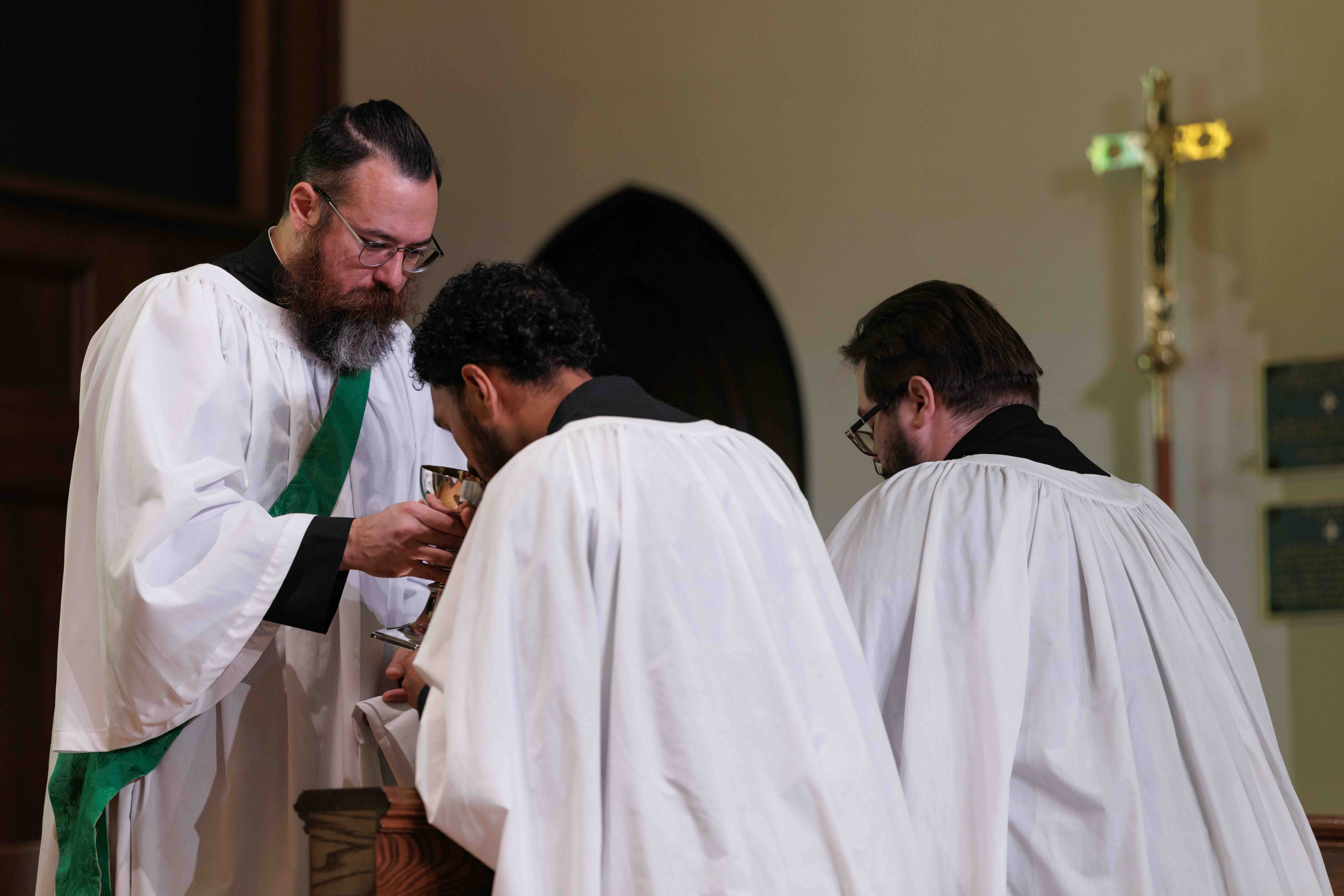 A priest, wearing a green stole over a white robe, offers communion to two men in white robes. A golden cross is visible in the background.