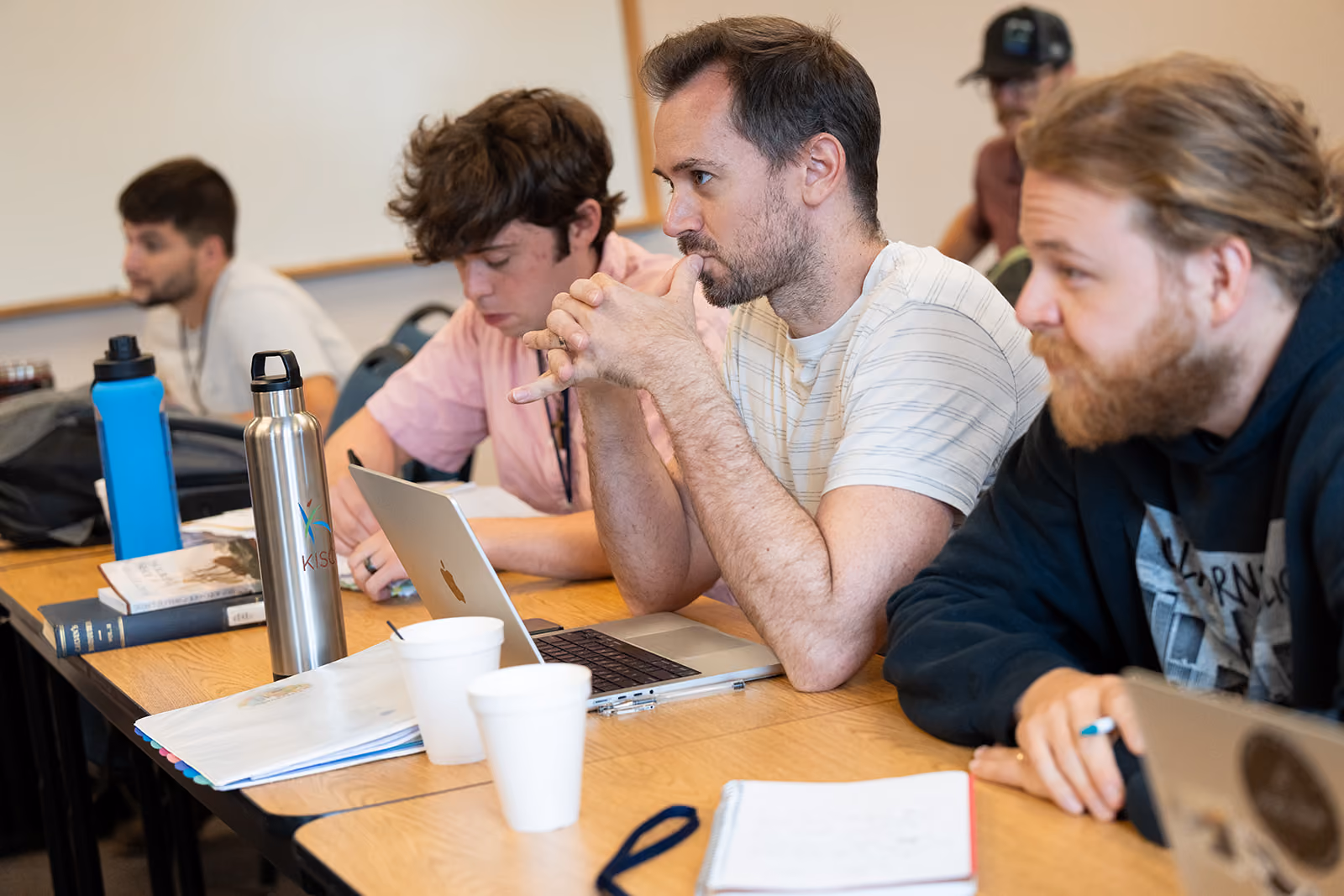 A group of four focused students sit at a classroom table with laptops, notebooks, and water bottles. They appear engaged in a discussion.
