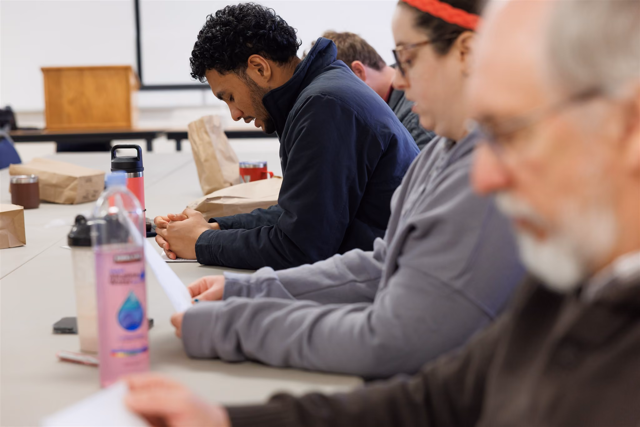 A group of people sit at a table, focused on papers in front of them. They have drinks and lunch bags nearby, creating a casual, thoughtful atmosphere.