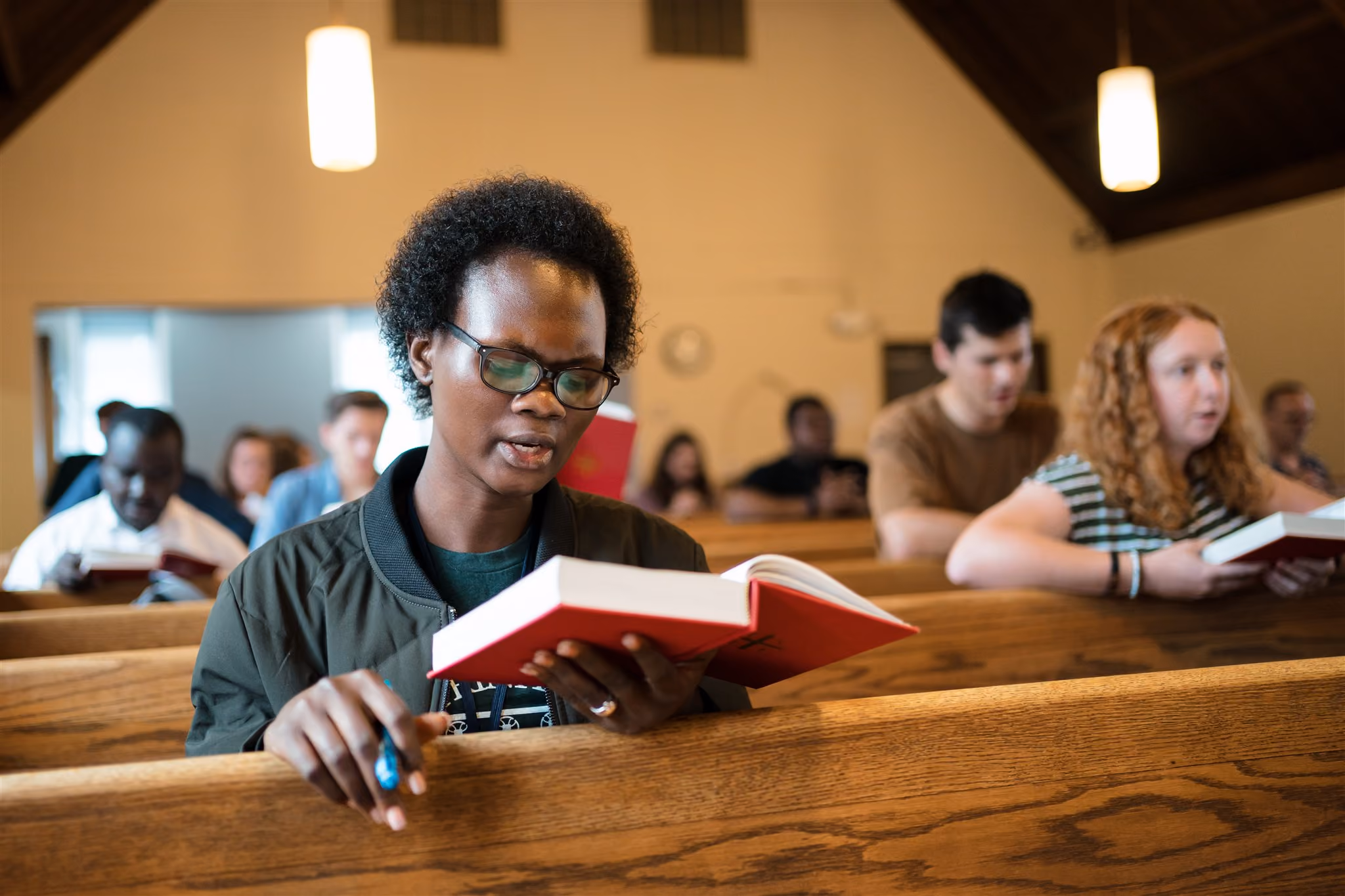 A diverse group of people sit in wooden pews, reading red hymnals in a church. The atmosphere is focused and serene, with soft lighting overhead.