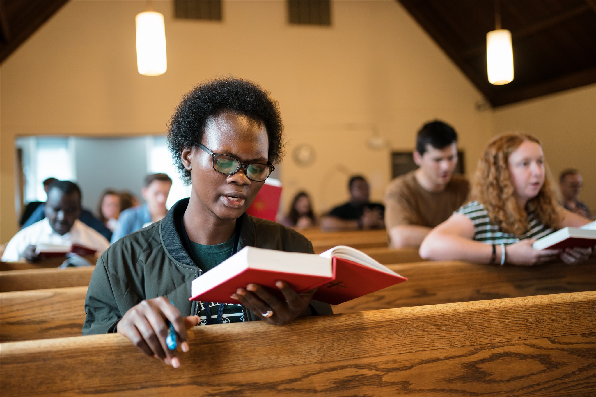 A diverse group of people sit in wooden pews, reading red hymnals in a church. The atmosphere is focused and serene, with soft lighting overhead.