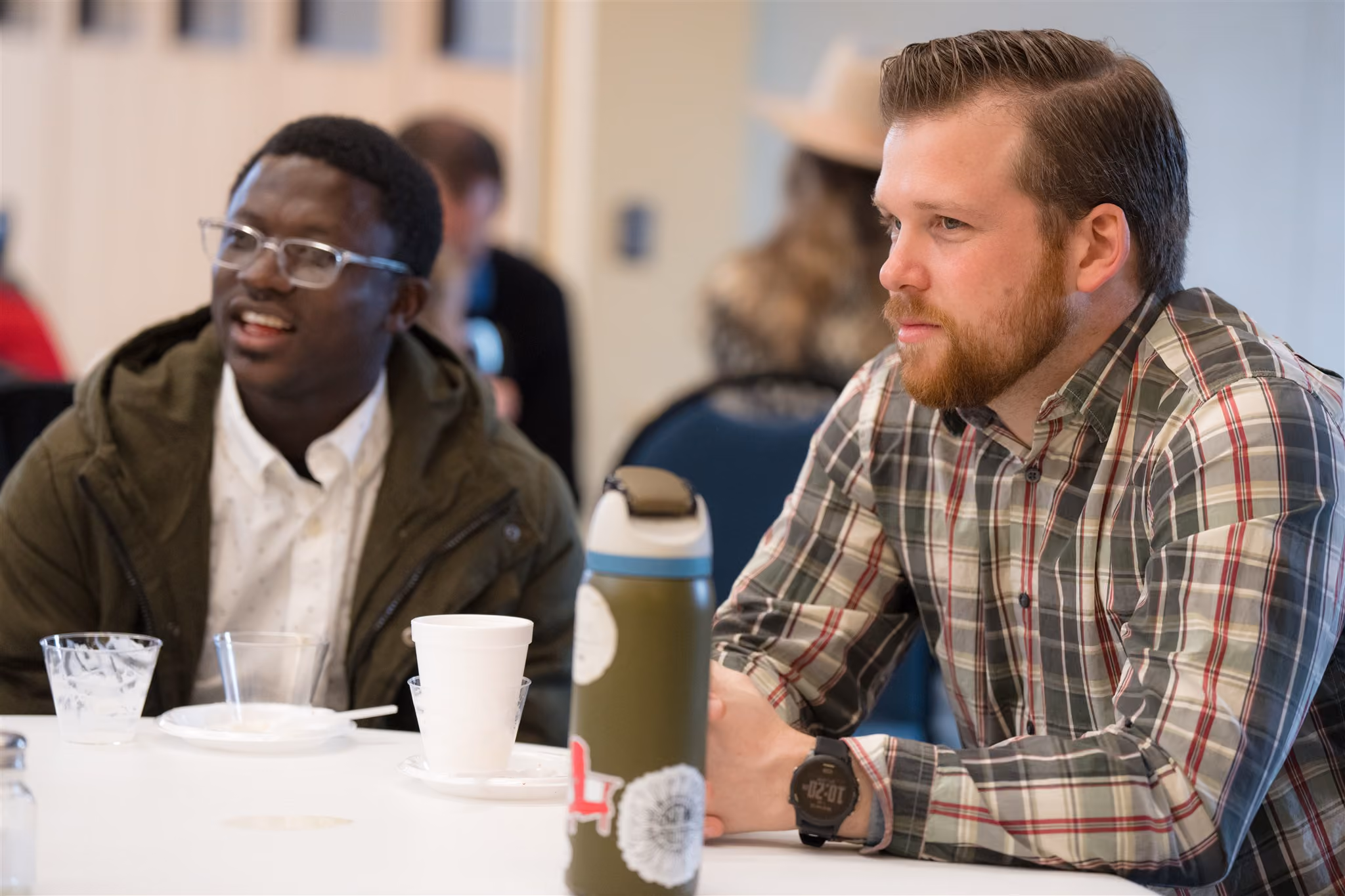 Two men sit at a table, engaged in conversation. One wears glasses and a green jacket, the other a plaid shirt. The setting is casual and friendly.