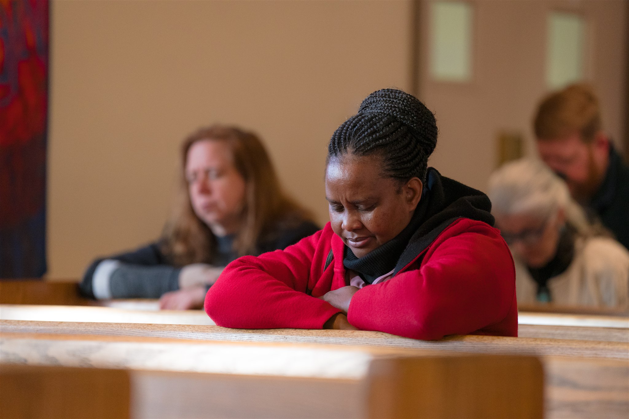 A woman in a red jacket, with eyes closed, leans forward on a church pew in prayer. Others in the background are also praying, creating a serene atmosphere.