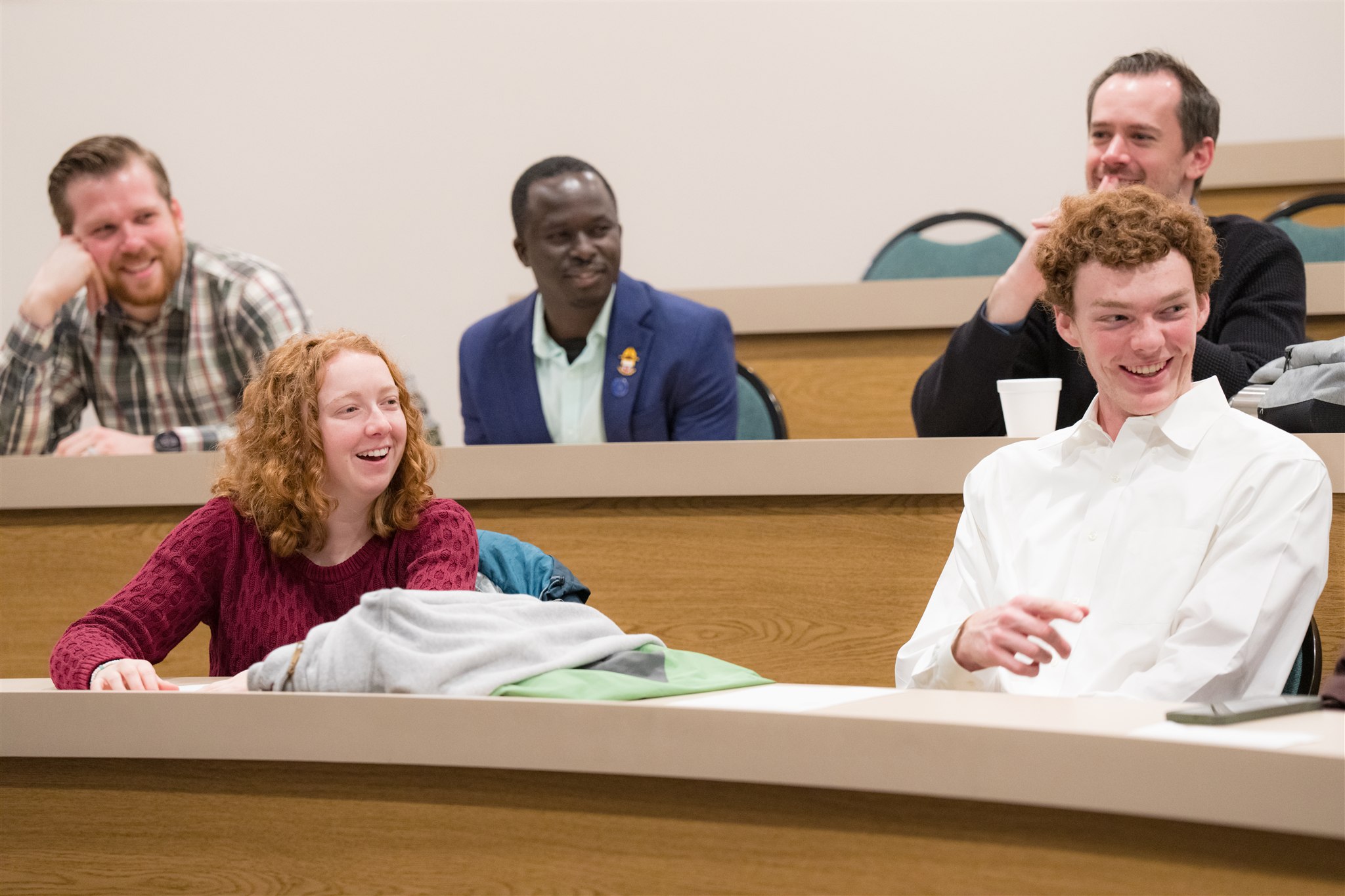 A diverse group of five people sit in a lecture hall, smiling and engaged, suggesting a lively and positive atmosphere. Classroom, interaction.