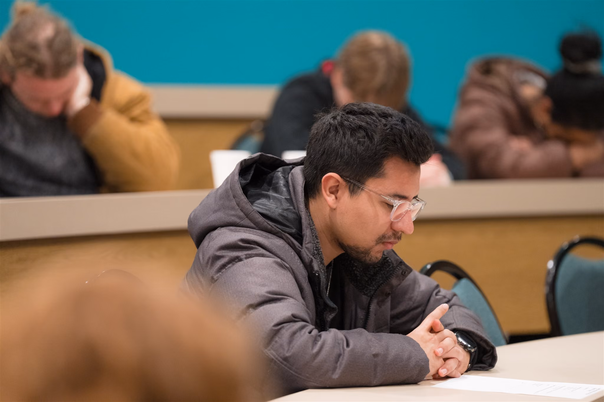 A group of people sits in a lecture hall with heads bowed in concentration. The mood is focused and serious, highlighting intense contemplation.