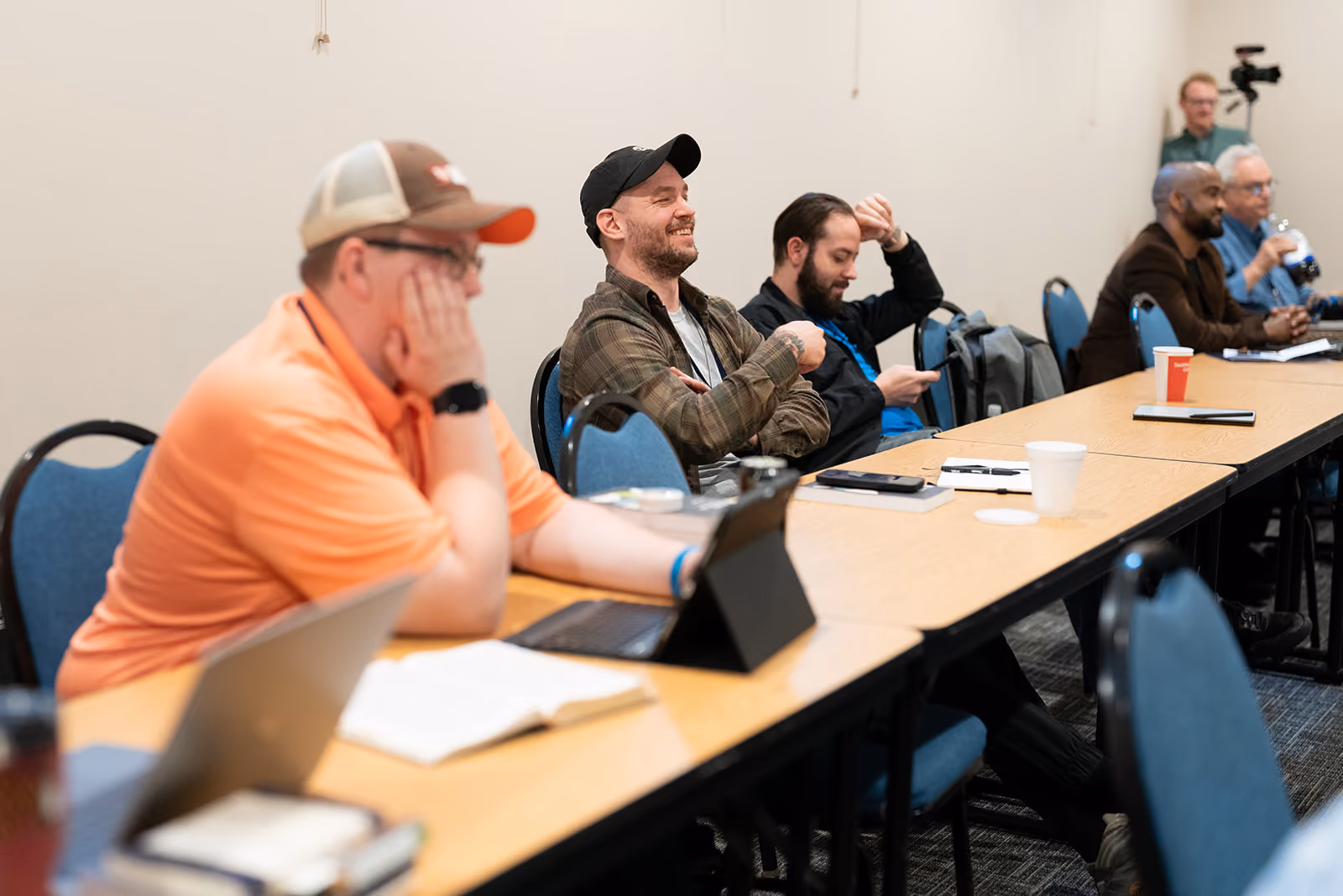 A group of people sit at tables in a classroom setting, some smiling and engaged, with notebooks and devices. The atmosphere is relaxed and attentive.