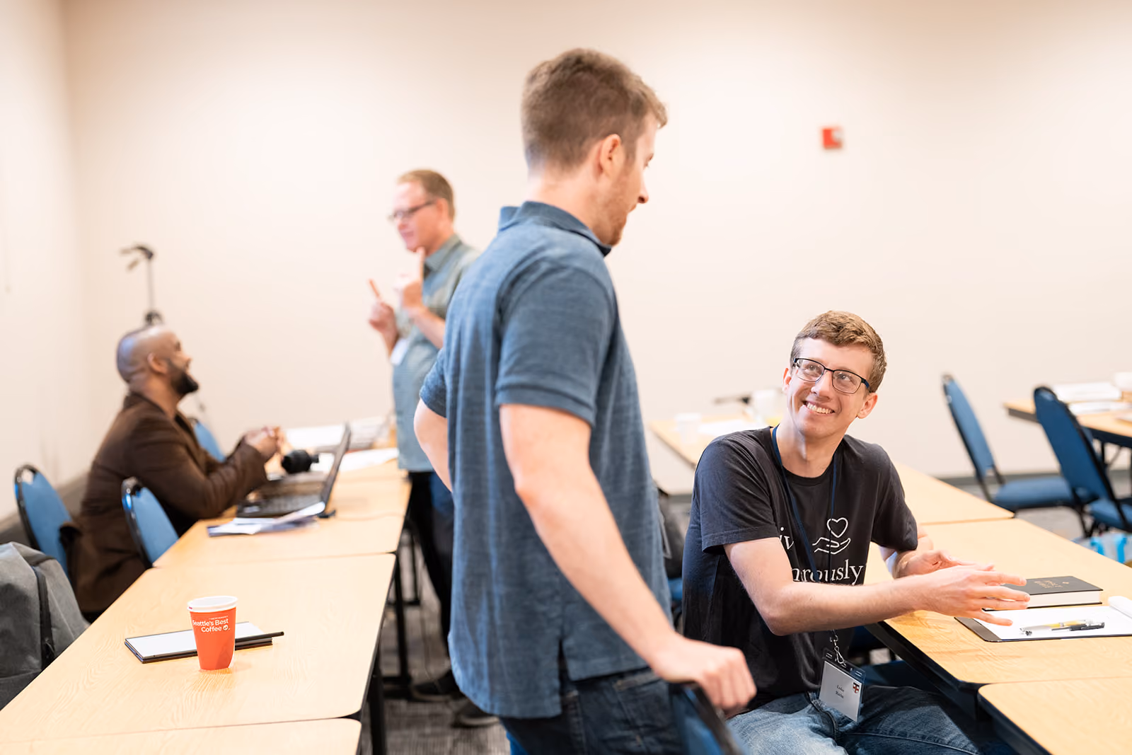 Two men engage in conversation in a classroom setting. One is seated, smiling up at the other. In the background, two more men chat. The atmosphere is friendly and engaged.