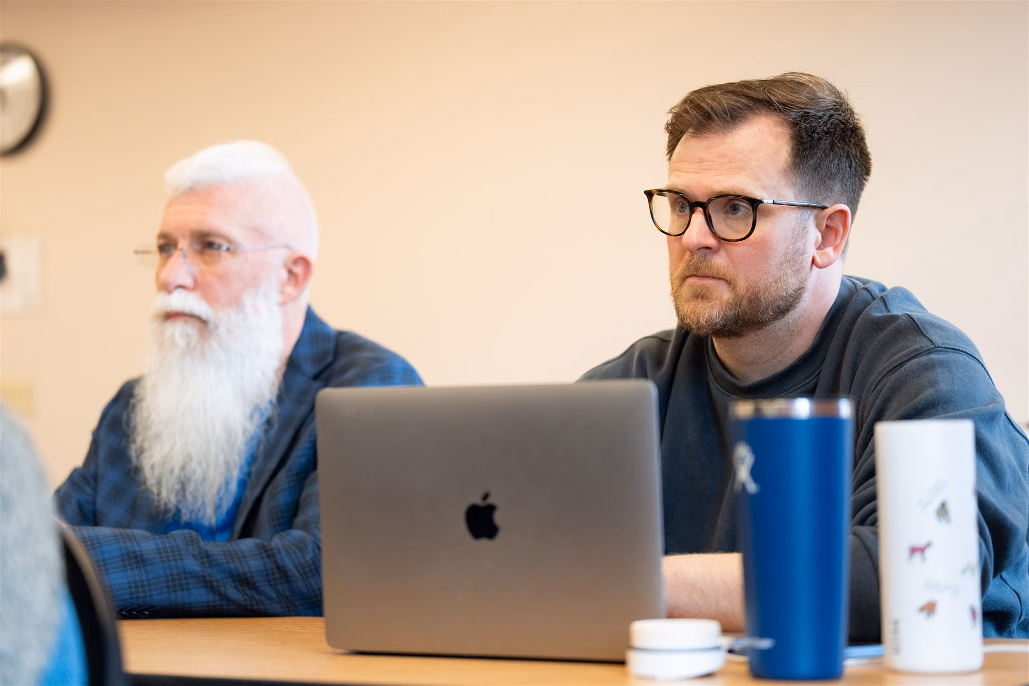 Two men, focused, sit at a table. One has a long white beard; the other wears glasses. A laptop, blue tumbler, and cup are in front of them.