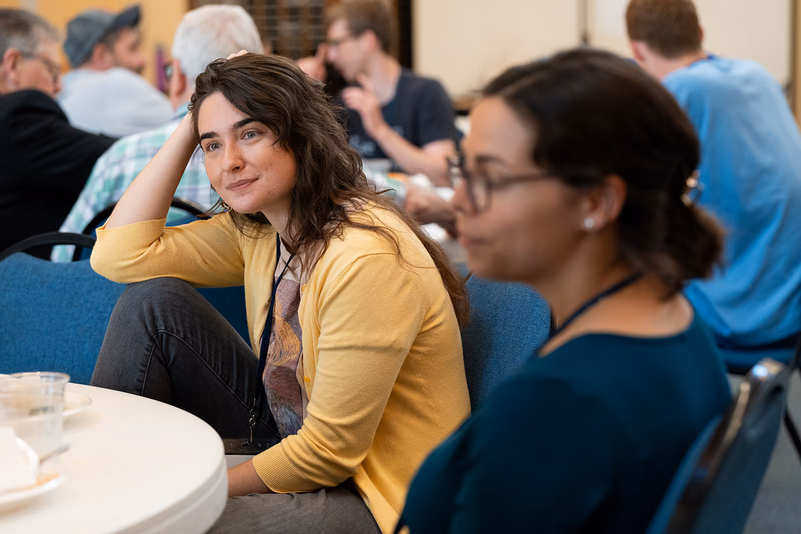 Two women seated in a conference room, engaged in conversation. One in a yellow cardigan appears attentive and thoughtful. People in the background.