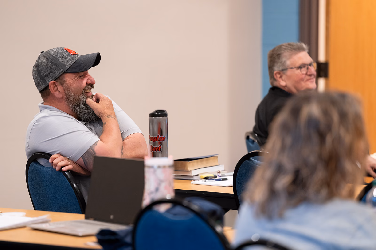 Two men sit in a classroom, one in a gray cap laughing. Books and a tumbler are on tables. The atmosphere is relaxed and engaging.