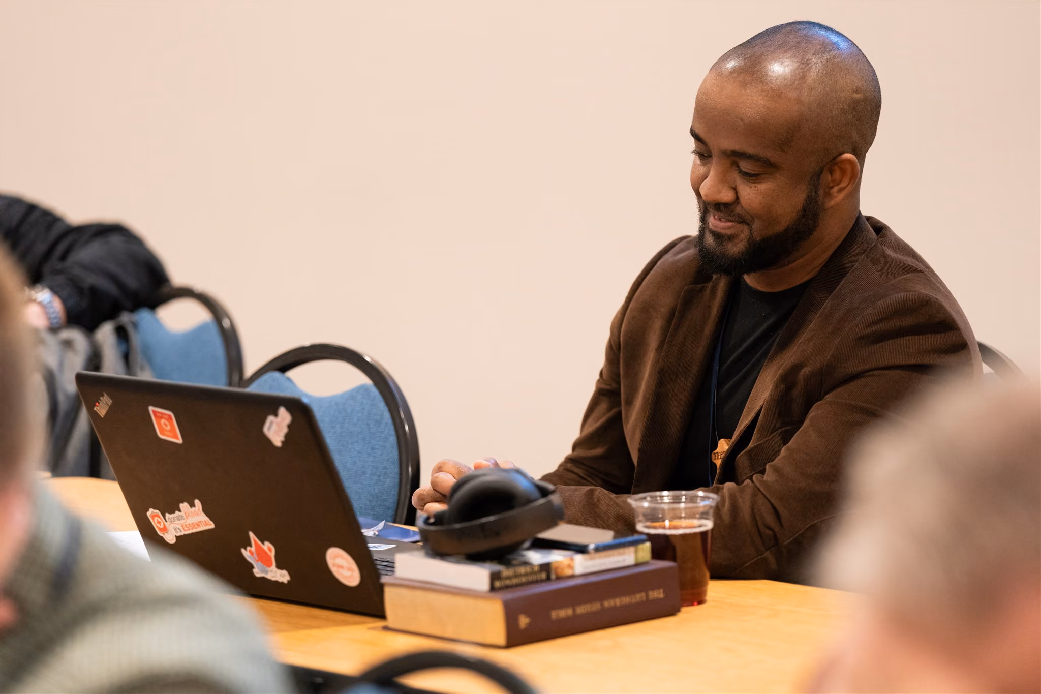 A man in a brown jacket, smiling and seated at a desk with a laptop covered in stickers, a stack of books, headphones, and a drink in a cup, creating a focused and relaxed atmosphere.