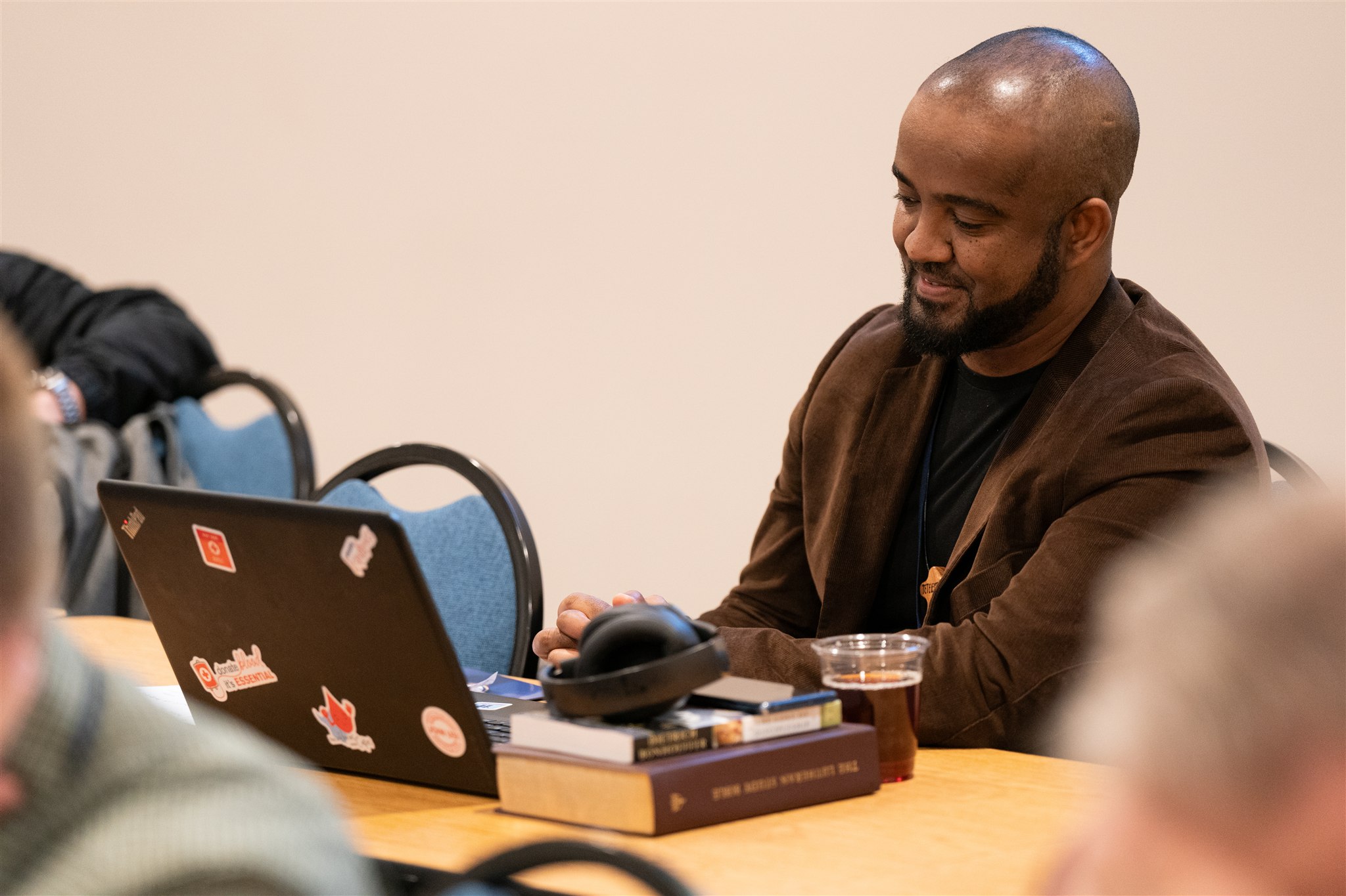 A man in a brown jacket, smiling and seated at a desk with a laptop covered in stickers, a stack of books, headphones, and a drink in a cup, creating a focused and relaxed atmosphere.