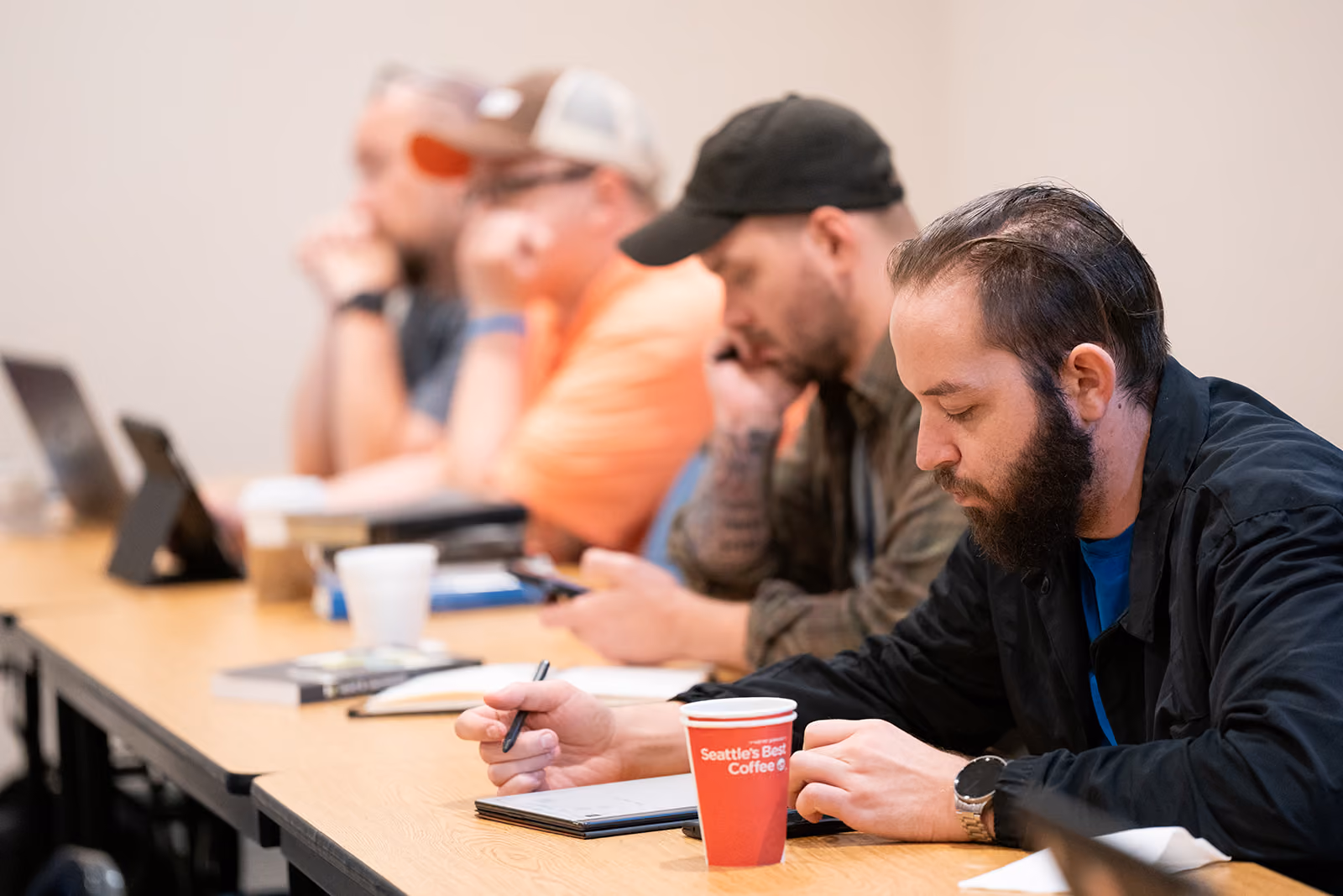 A group of men attentively seated at a conference room table, with notebooks and coffee cups. The atmosphere is focused and contemplative.