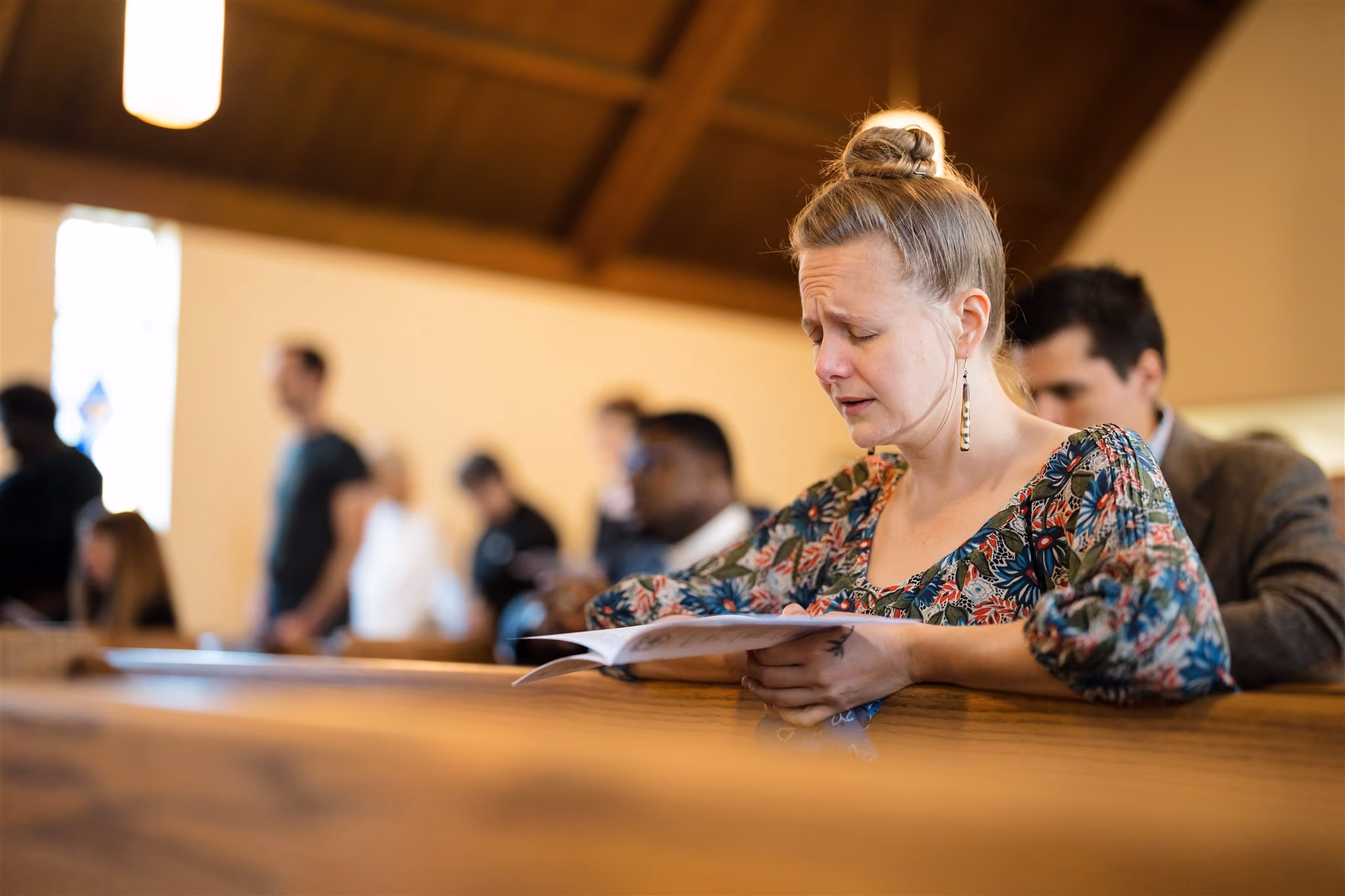 A woman sits in a church pew, eyes closed and appearing emotional, holding a book. Other people are in the background, some standing, in soft focus.