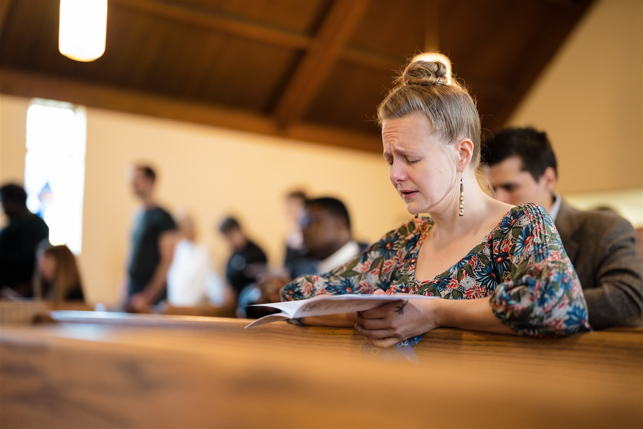A woman sits in a church pew, eyes closed and appearing emotional, holding a book. Other people are in the background, some standing, in soft focus.