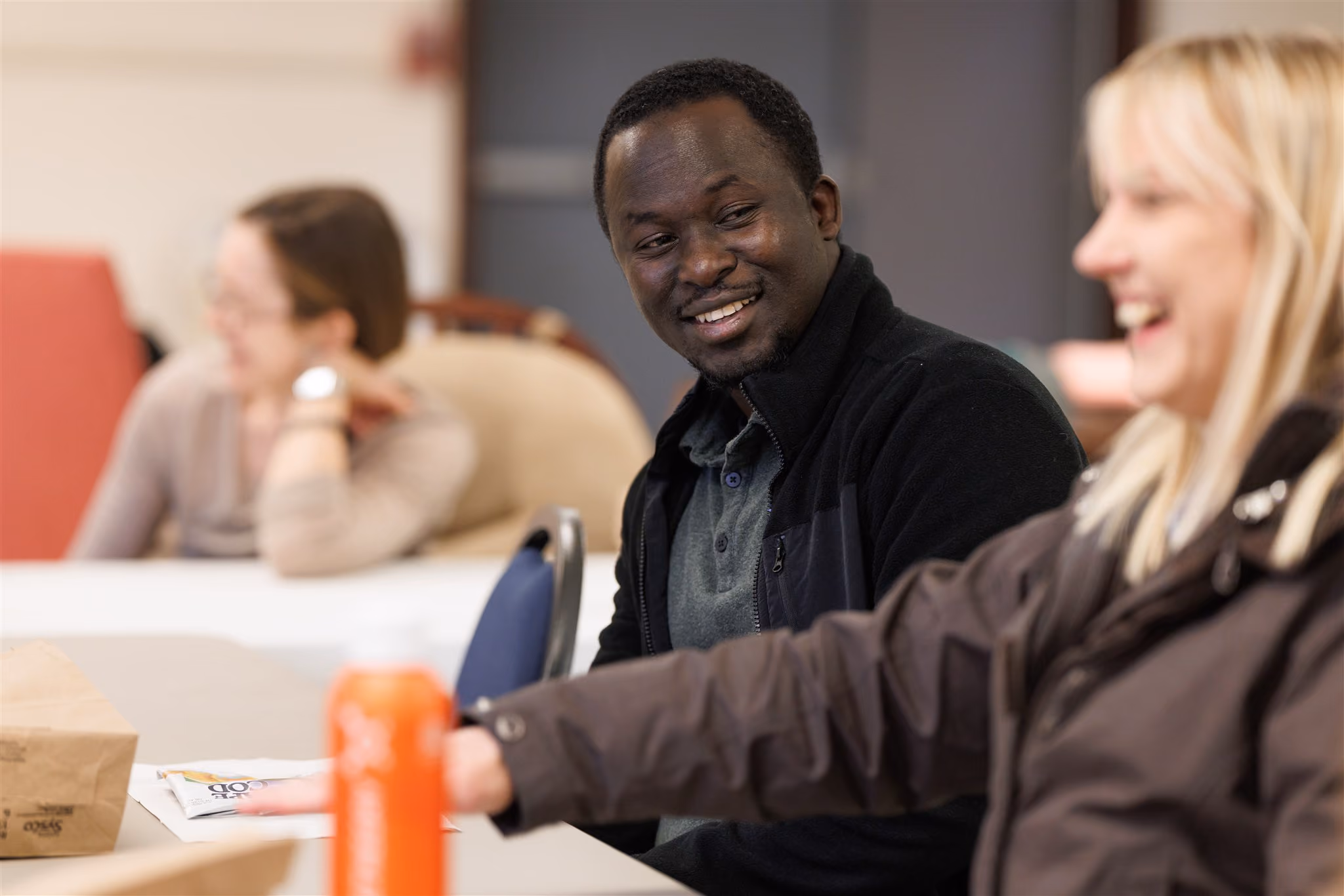 A group of people sit at a table, engaging in a lively conversation. The foreground shows a man smiling at a laughing woman, conveying a joyful atmosphere.