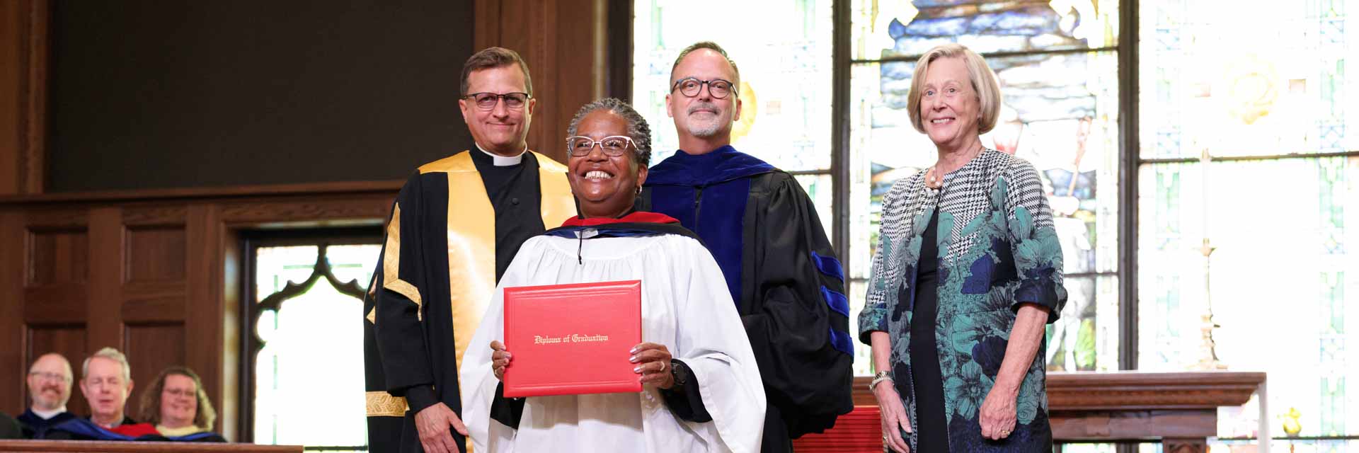 A woman in graduation attire is being hooded by a man in academic robes. They stand in a church-like setting, conveying a sense of achievement.