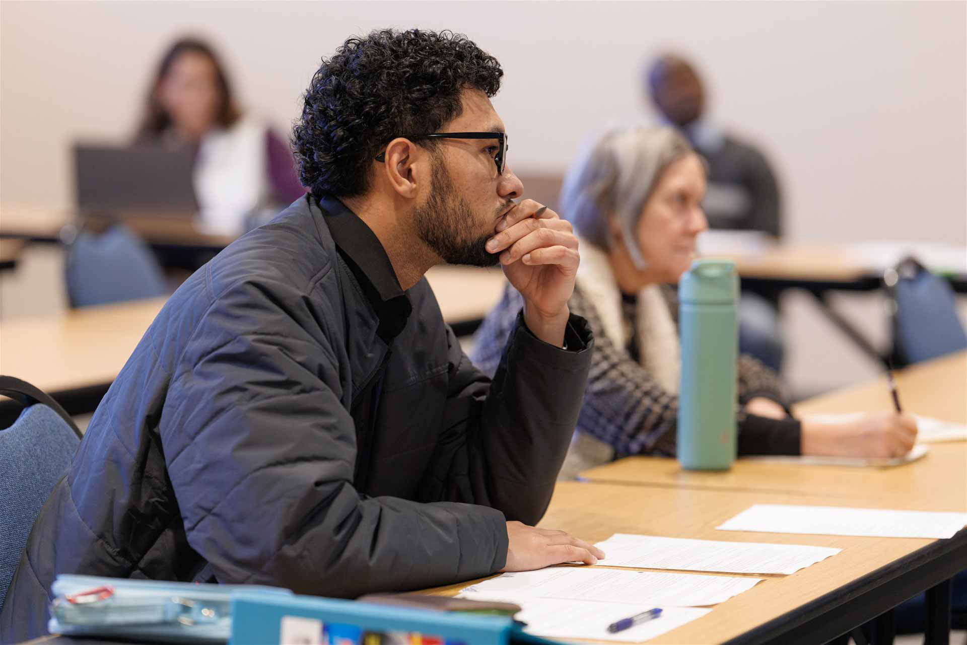A thoughtful man in glasses and a jacket sits in a classroom, chin resting on hand. Focused, he listens intently, papers and a water bottle nearby.