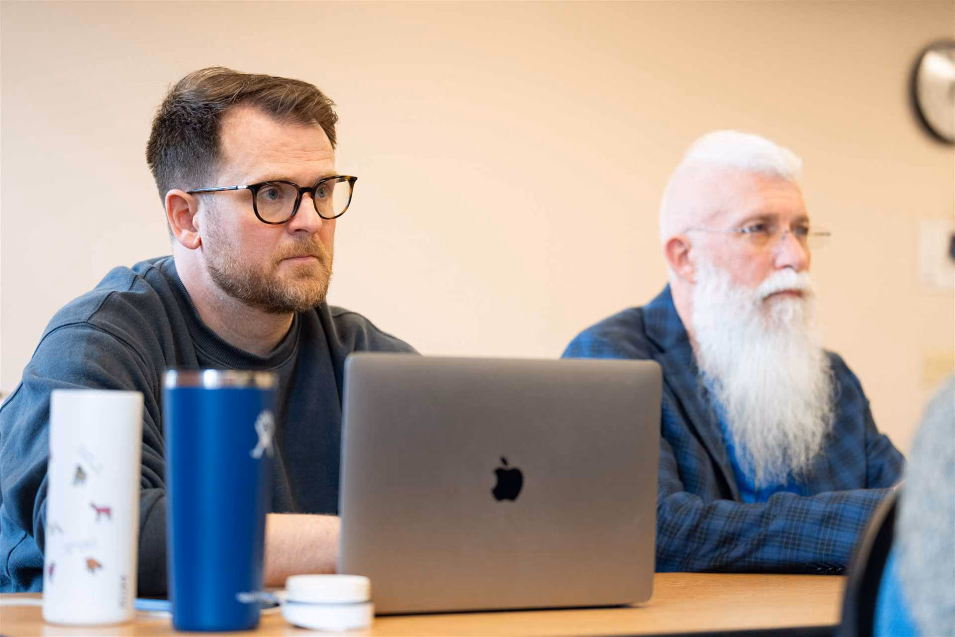 Two men sitting at a table, focused on a meeting. One has a white beard and glasses; the other wears glasses and sits behind a laptop.