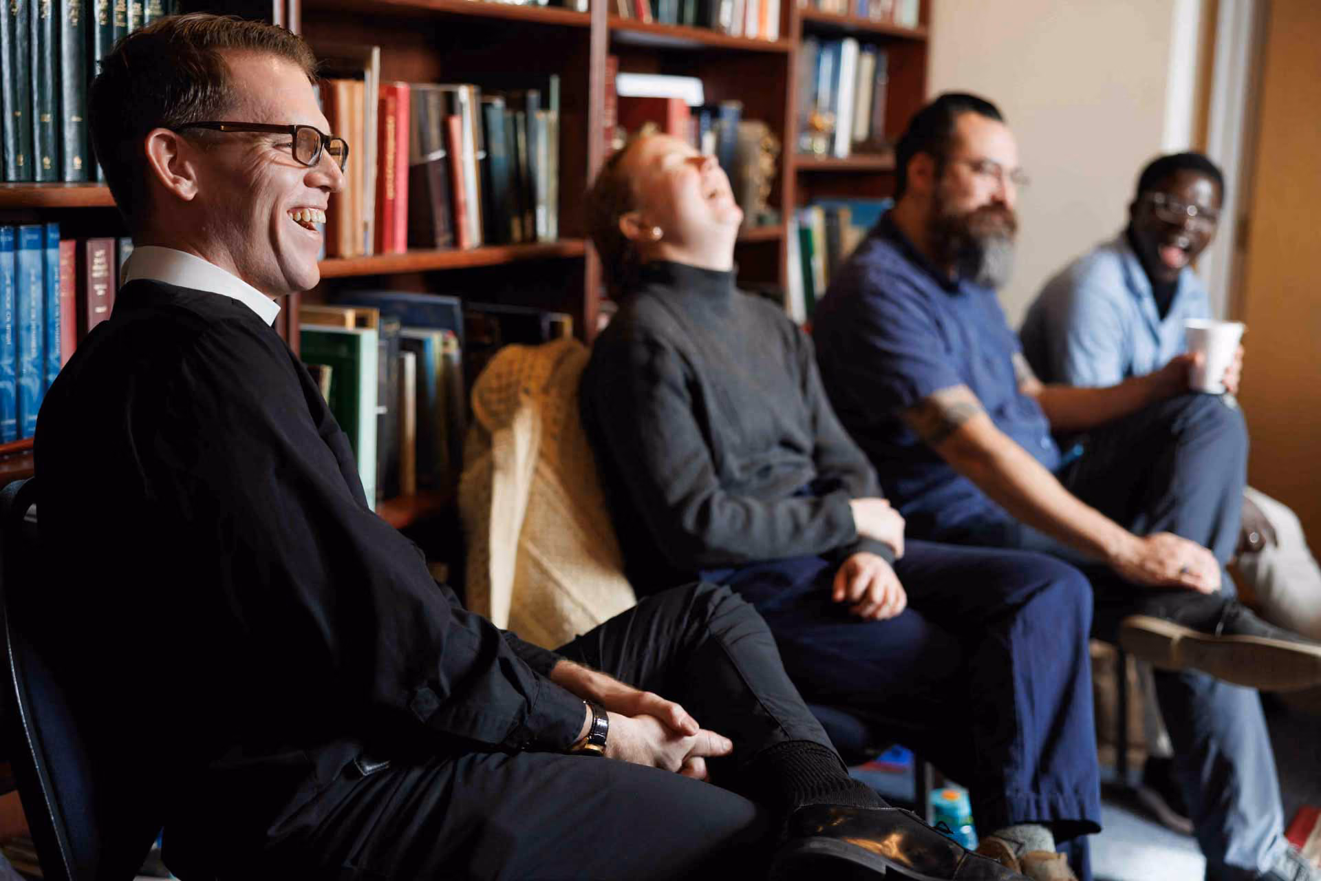 A group of four people sits in a library, laughing and smiling. Books fill the shelves behind them, creating a warm, relaxed atmosphere.