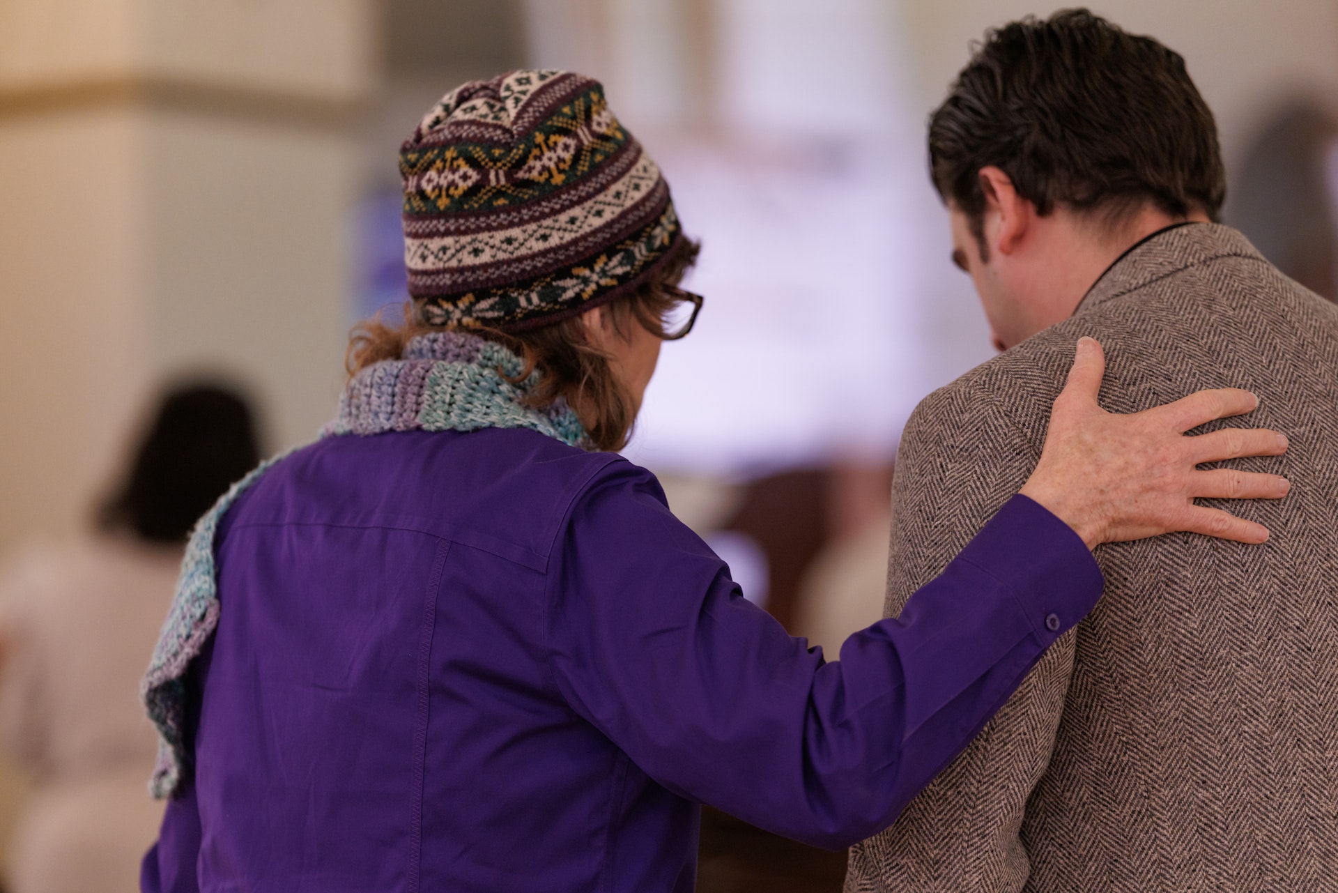 A person in a purple jacket and patterned hat gently places a hand on the shoulder of a man in a tweed coat, conveying comfort and support indoors.