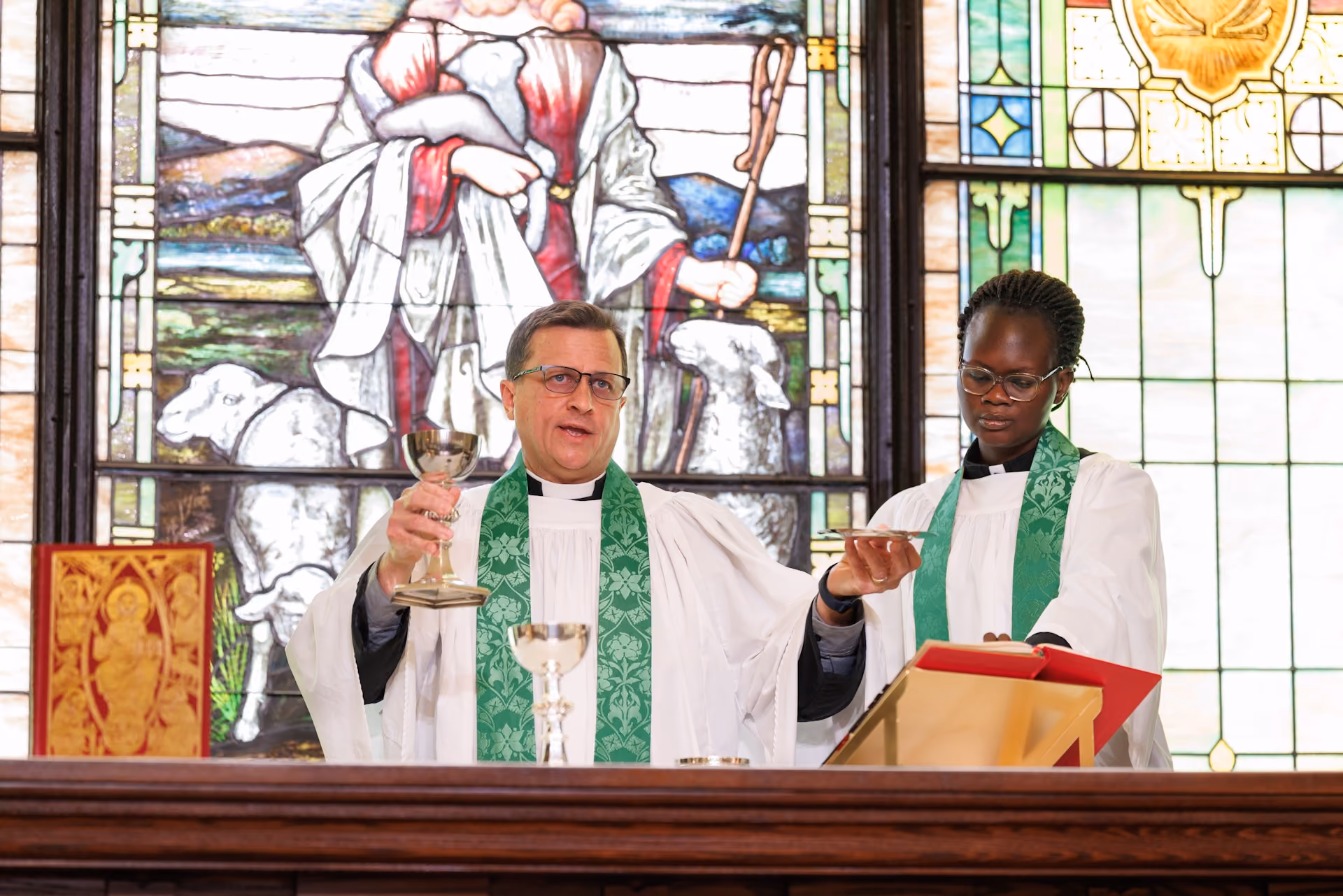 Two clergy in white robes with green stoles stand at an altar holding a chalice and book, against a colorful stained glass backdrop depicting religious imagery.