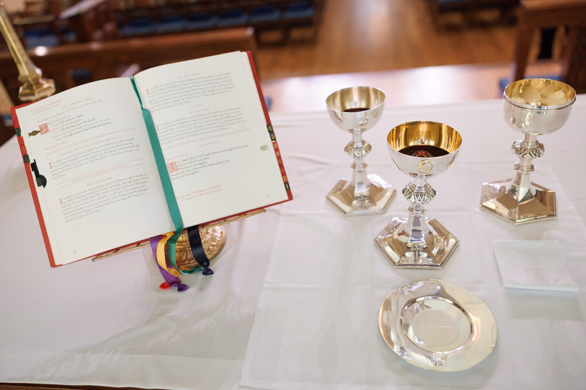 An altar with an open liturgical book, three golden chalices, and a silver plate on a white cloth. The church interior is softly lit, evoking reverence.