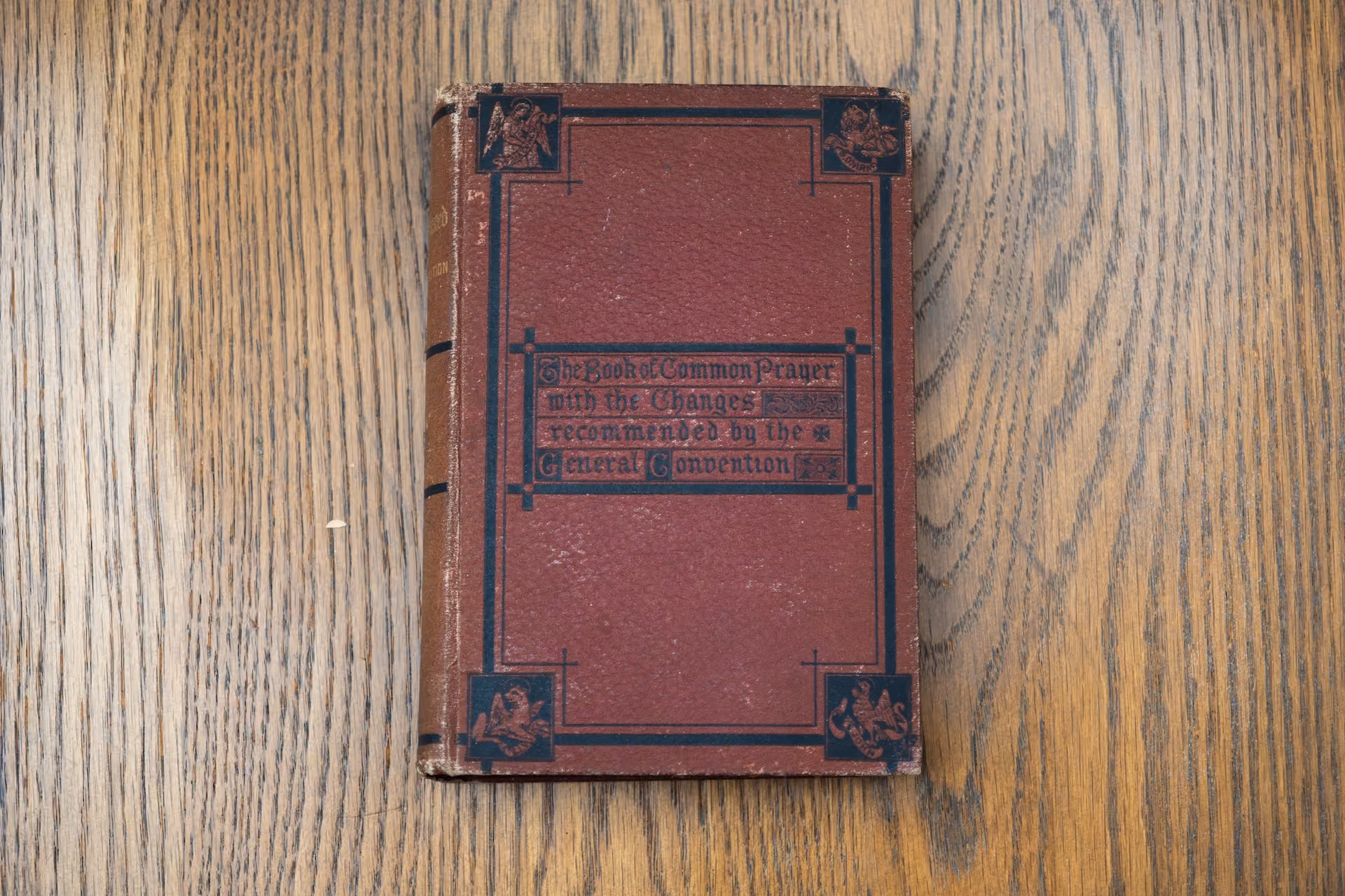 A vintage, red leather-bound book titled "The Book of Common Prayer" lies on a wooden surface. The cover features decorative black borders and corners.