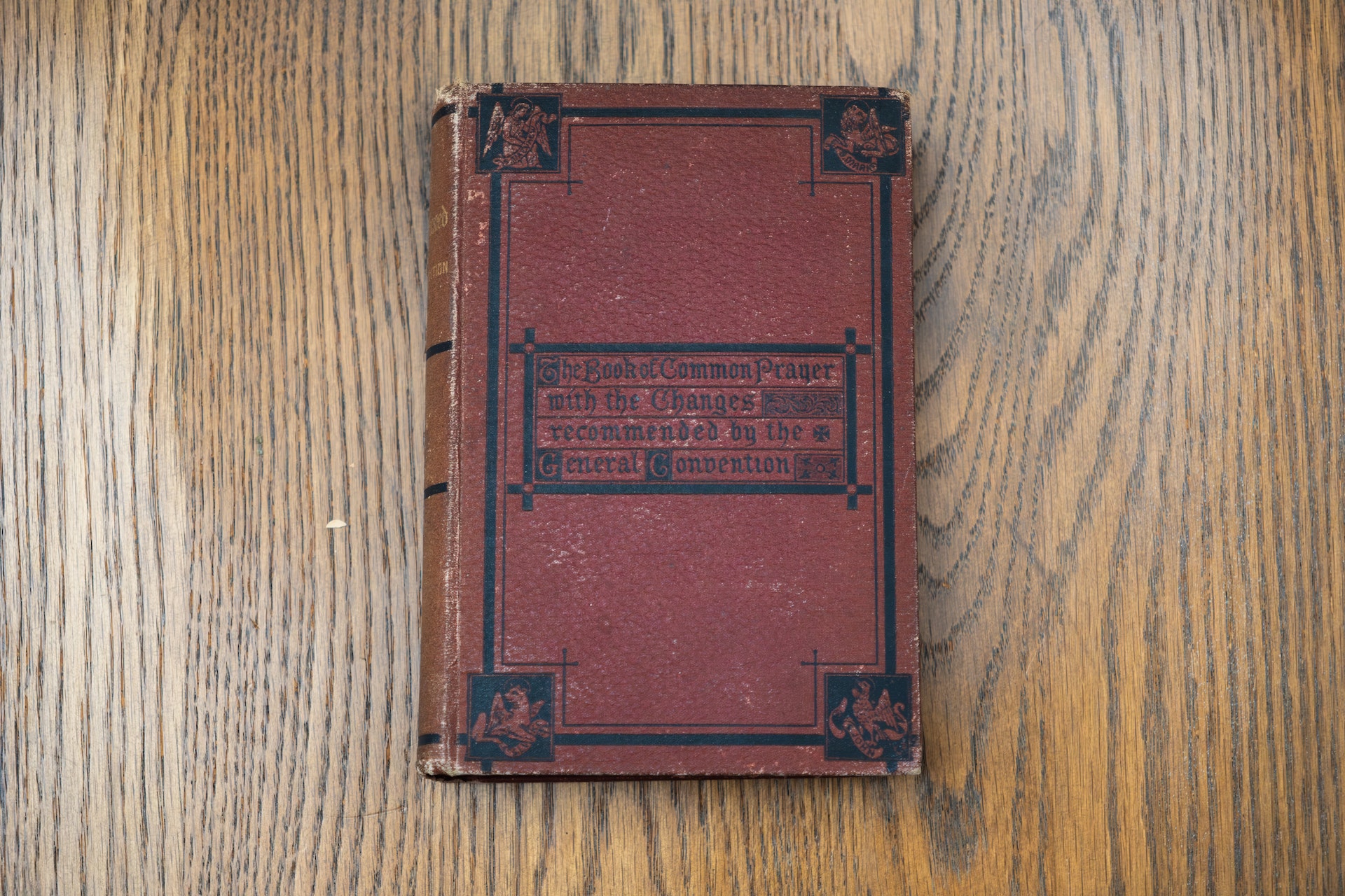 A vintage, red leather-bound book titled "The Book of Common Prayer" lies on a wooden surface. The cover features decorative black borders and corners.