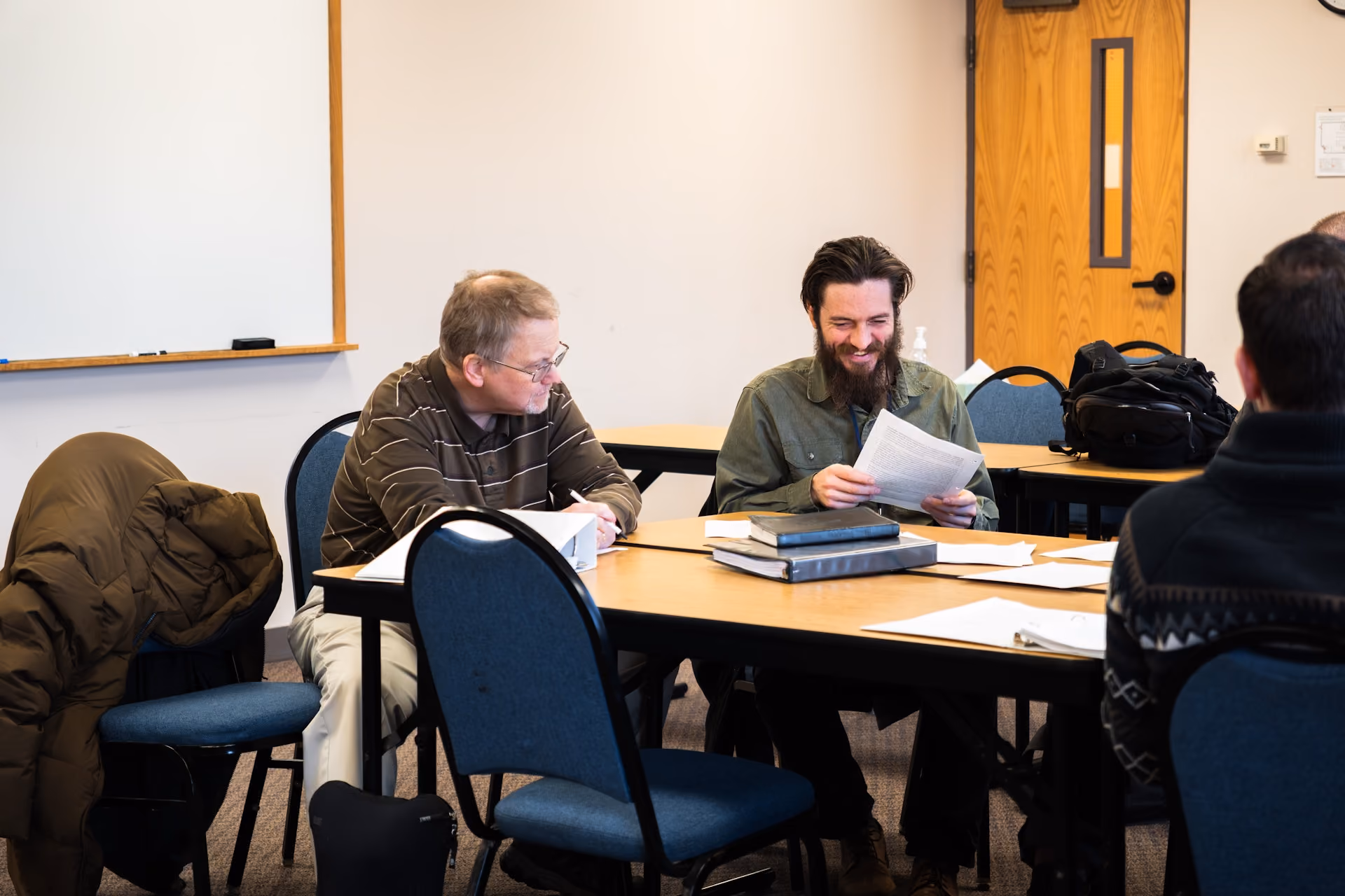 Two men sit at a classroom table, smiling and discussing papers. Books and jackets are on nearby chairs, conveying a relaxed learning atmosphere.