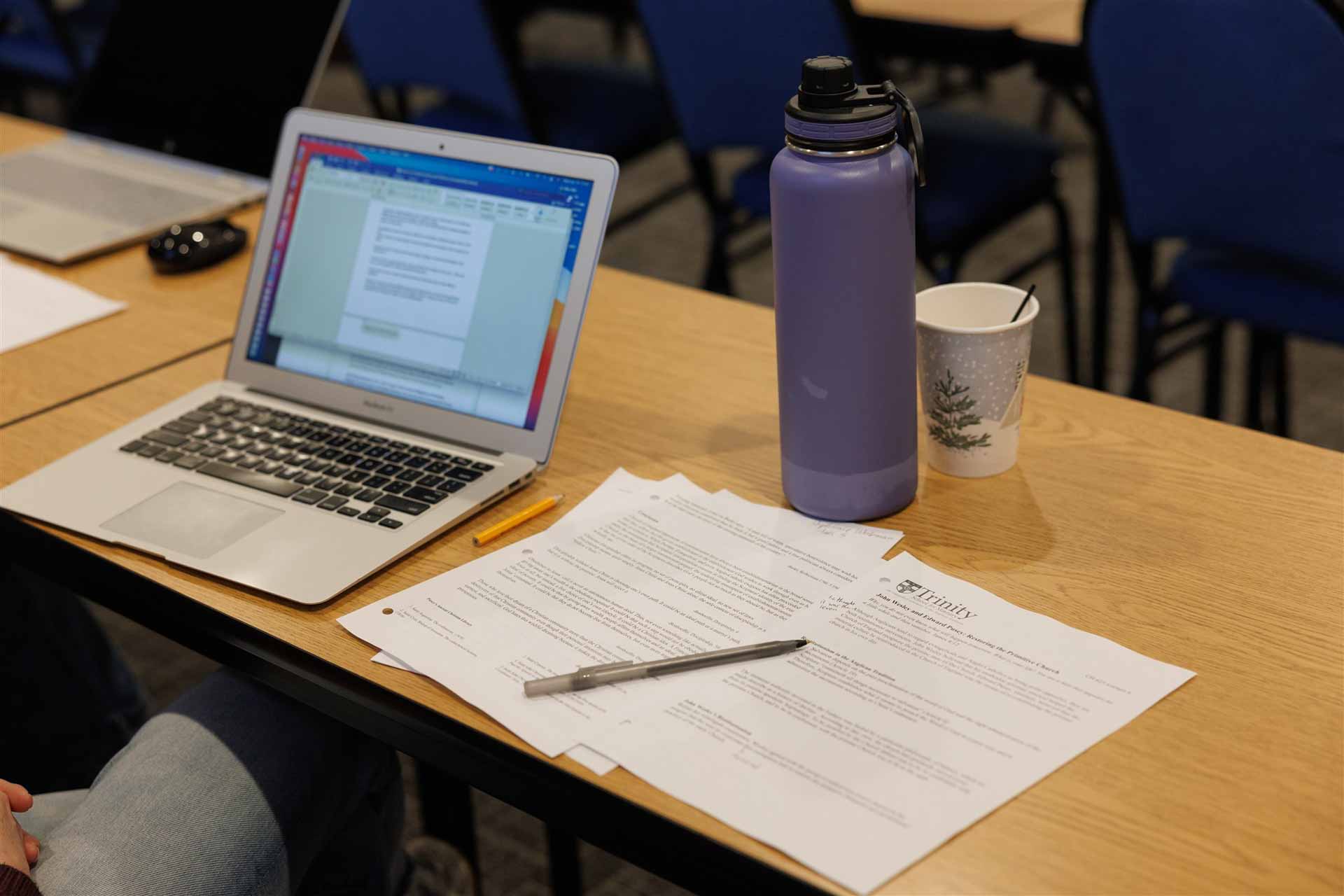 Open laptop showing a document on a wooden desk, alongside printed papers, a gray pen, a purple water bottle, and a paper cup; classroom setting.