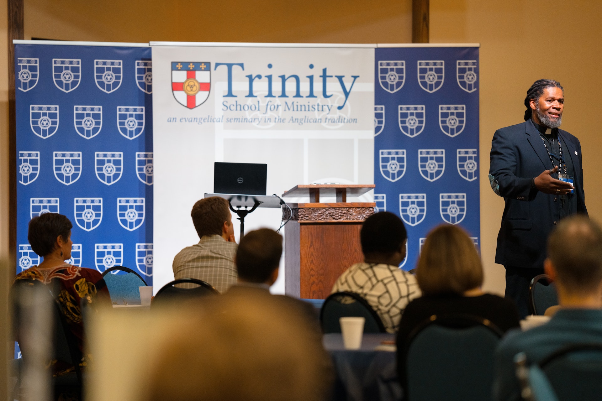 A speaker presents enthusiastically at a Trinity School for Ministry event, with attendees listening. The backdrop features the school's logo and branding.